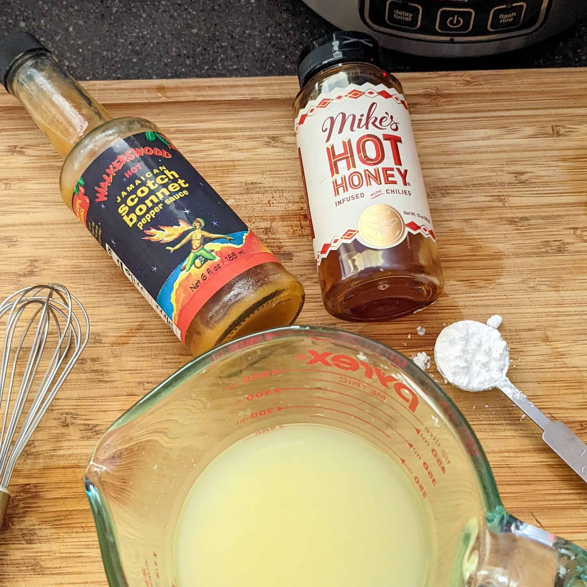 chicken broth in a glass measuring cup, next to mike's hot honey and walkerwood's scotch bonnet pepper sauce.