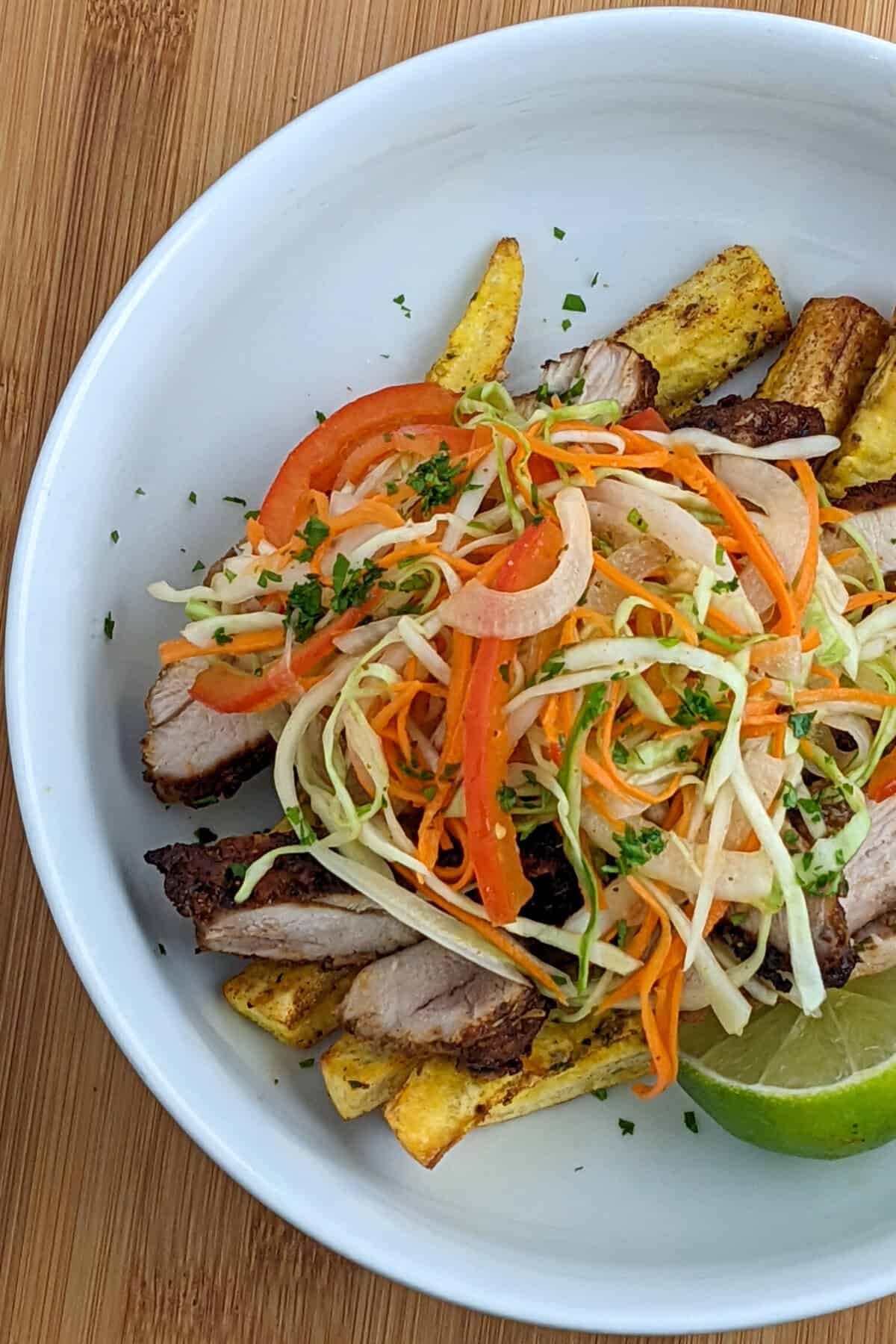 loaded plantain fries in a round dish with tall sides topped with sliced seasoned chicken thighs and pikliz (a hot pepper cabbage slaw).