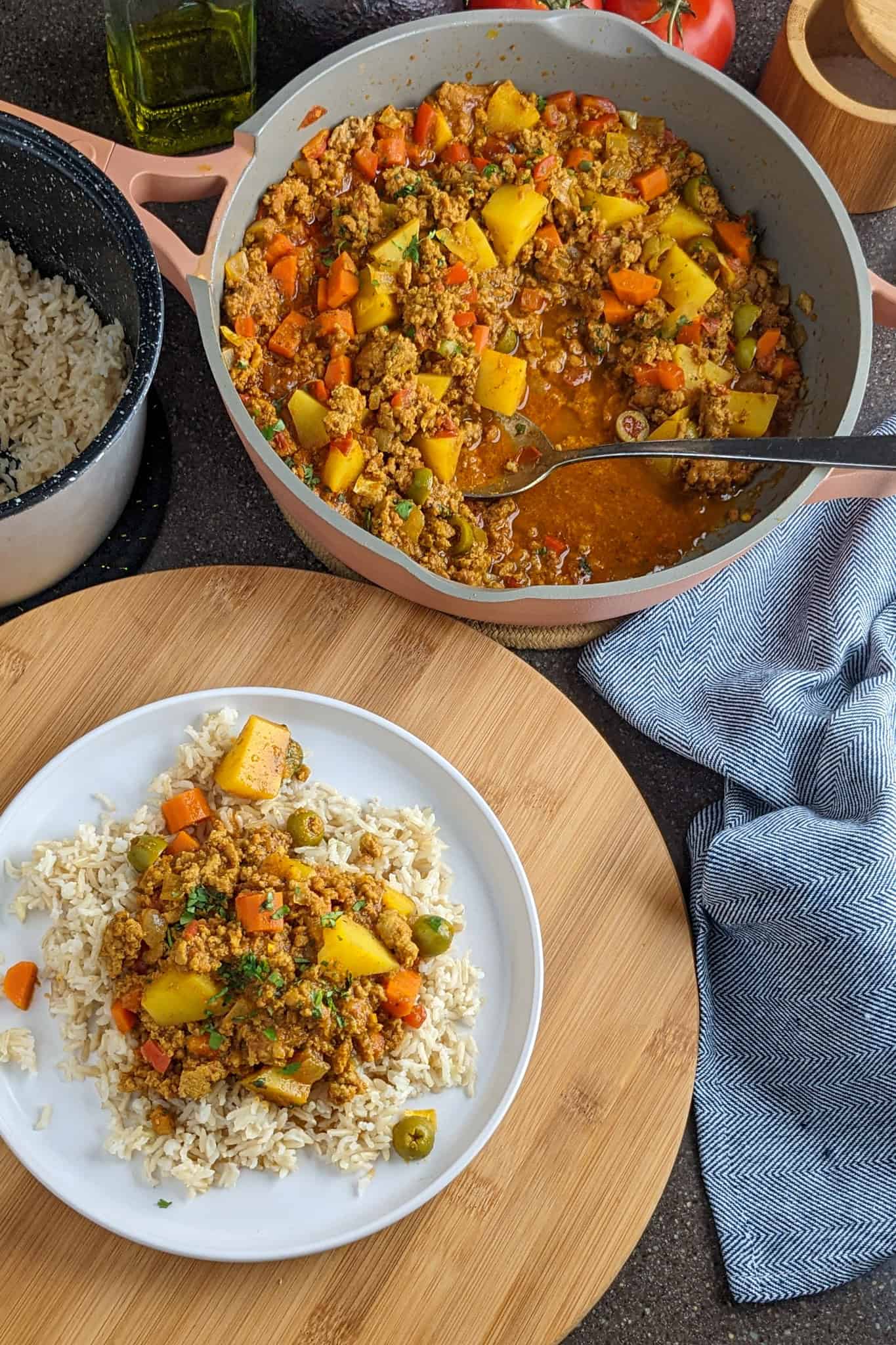 a plate of curried ground turkey picadillo over rice on a flat round plate surrounded by a pot of more picadillo and a canister of cooked rice.