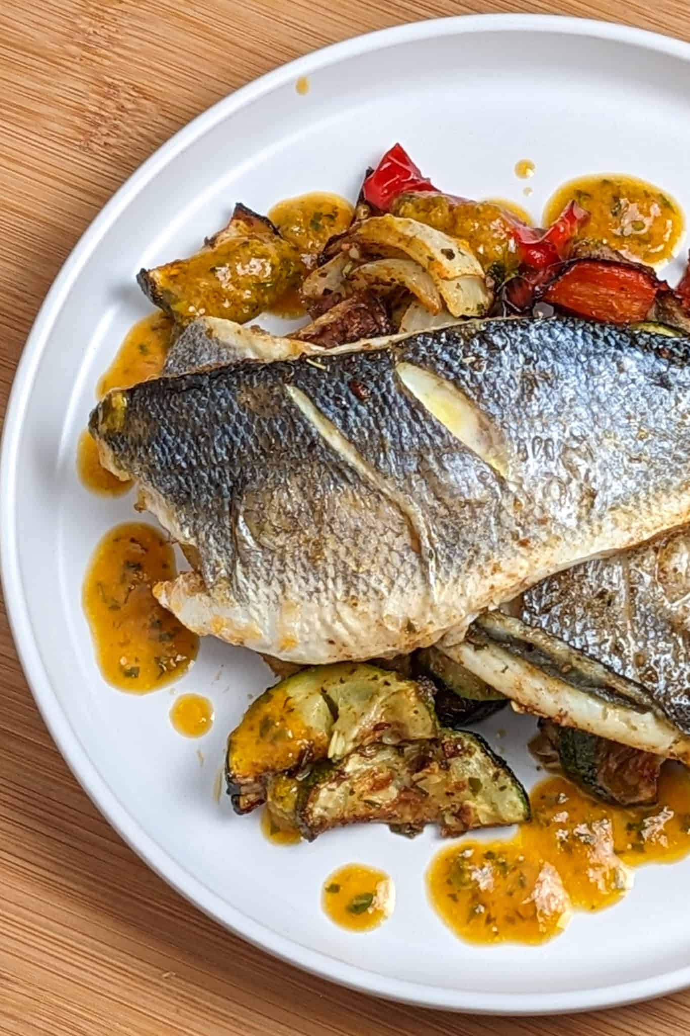 air fried branzino resting on cooked sliced vegetables surrounded by lemon sauce on a flat round plate.