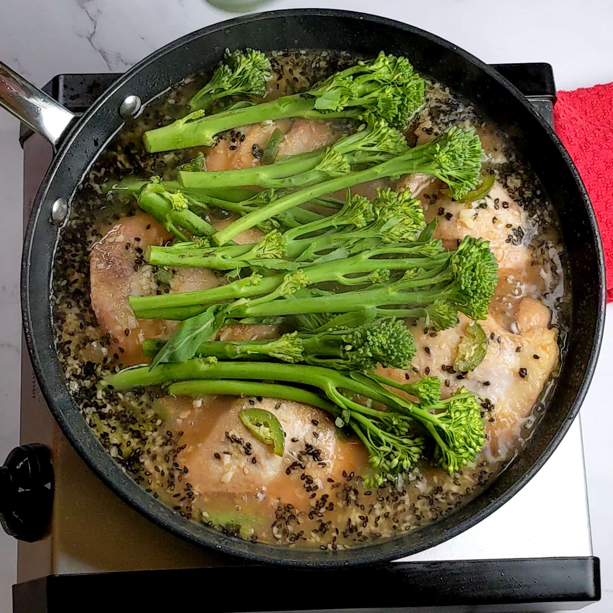 raw broccolini sitting on top of the seared sesame chicken in the seasoned broth in a non-stick frying pan