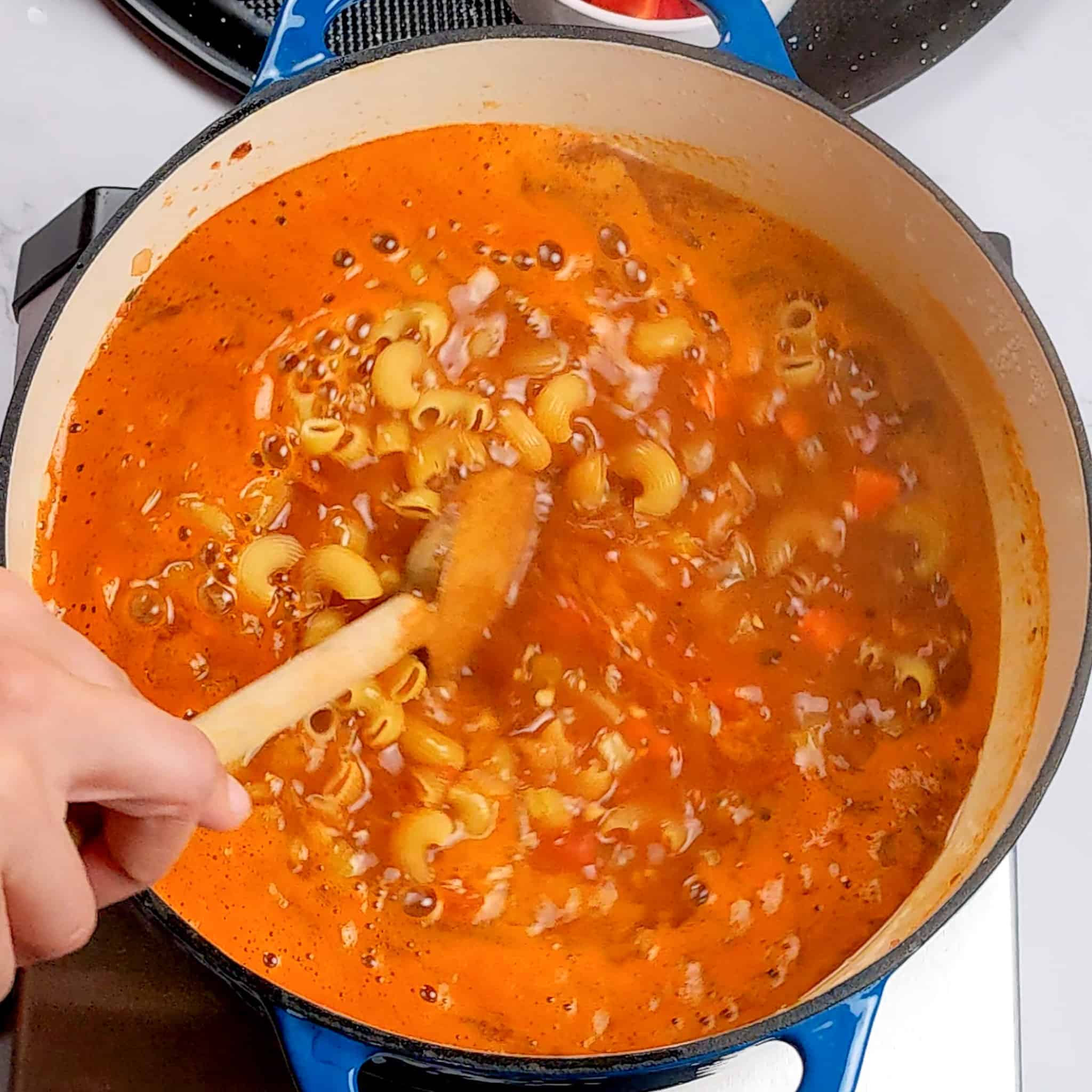 raw macaroni pasta being stirred in a rich tomato broth with a wooden spoon in a dutch oven