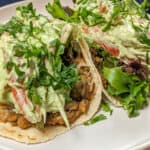 two Salsa Verde Turkey and Beans Tacos with Avocado Cream on a white round plate with shallow sides on a wooden lazy susan with a kitchen towel in the background.