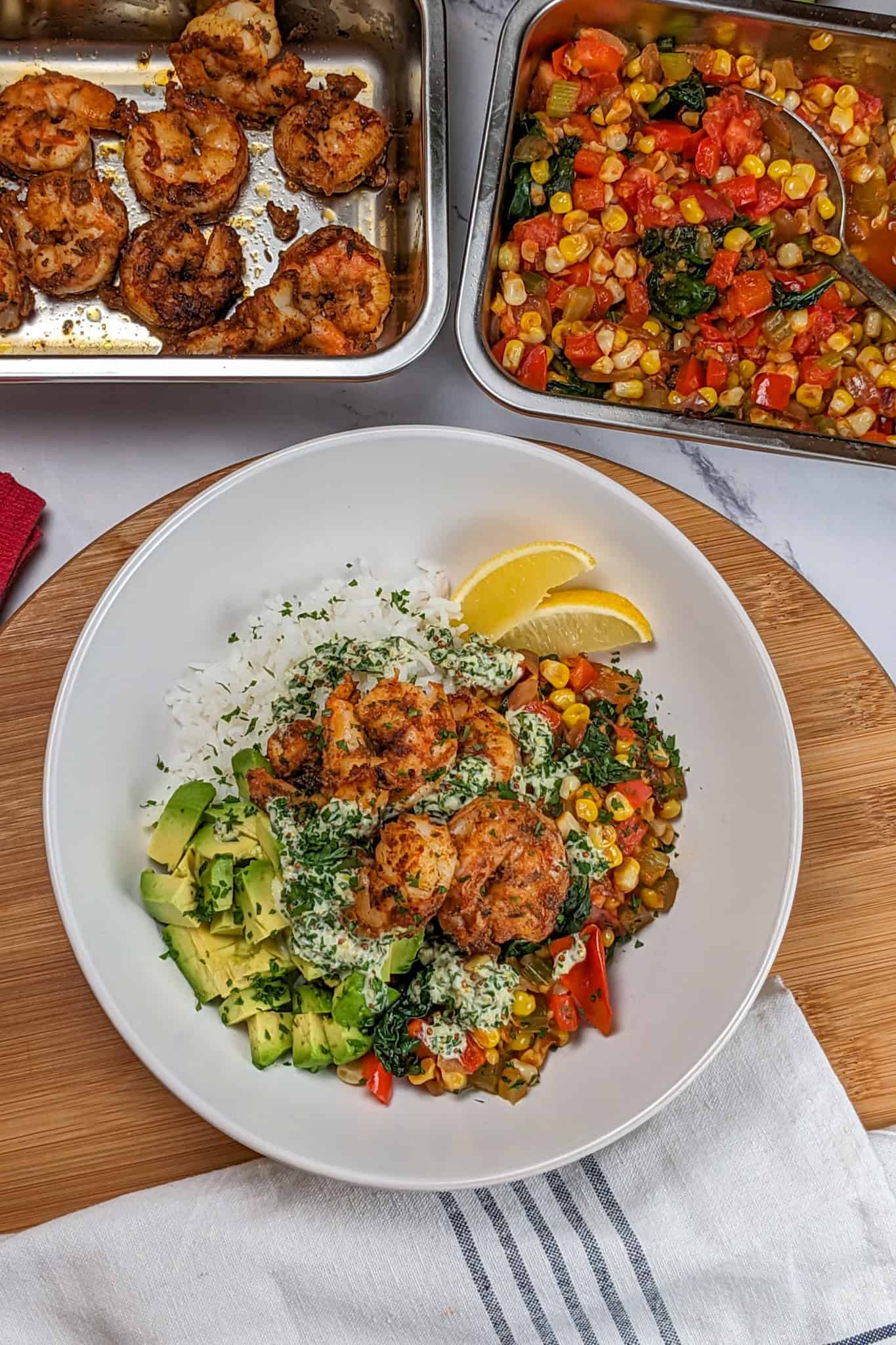 Top view zoomed out picture of the Spicy Cajun Shrimp Rice Bowl with Lemon Remoulade in a wide rim bowl with diced avocado and lemon wedges, next to a kitchen towel and two rectangle metal dishes with more cajun shrimp and vegetables.