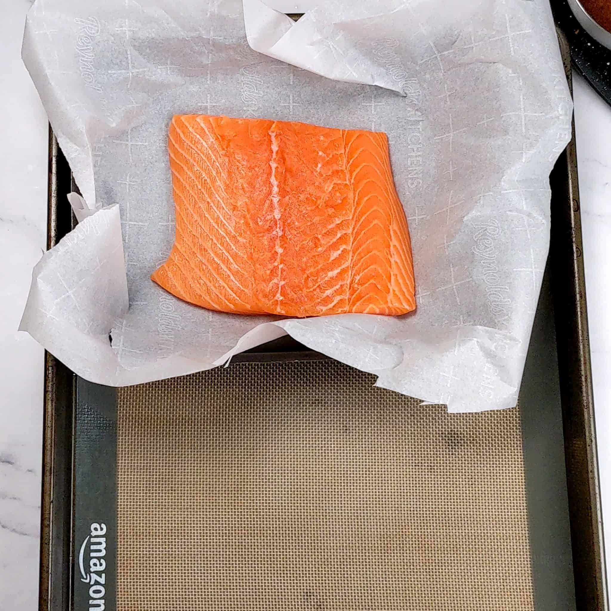 salmon filet on a parchment paper lined baking dish resting on a silicone lined sheet pan.
