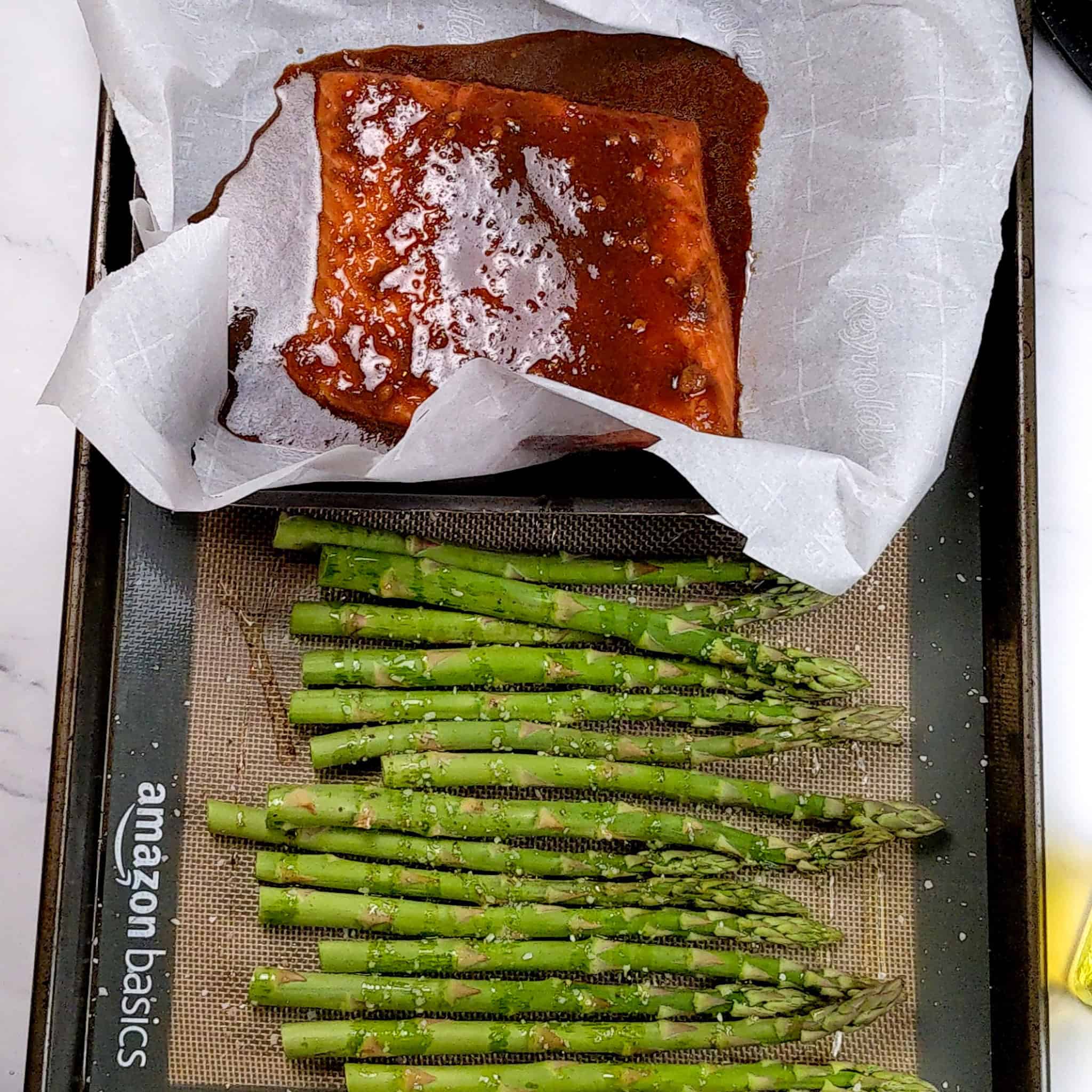 raw salmon filet drenched in the miso ginger sauce on a parchment paper lined baking dish resting on a silicone lined sheet pan with lined up asparagus seasoned with oil, salt and pepper.