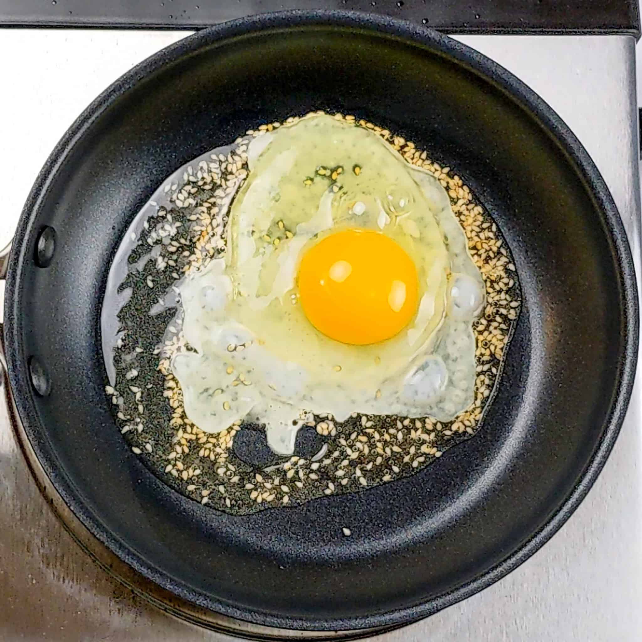 cracked egg with sesame seeds frying in oil in a non stick oxo pan.
