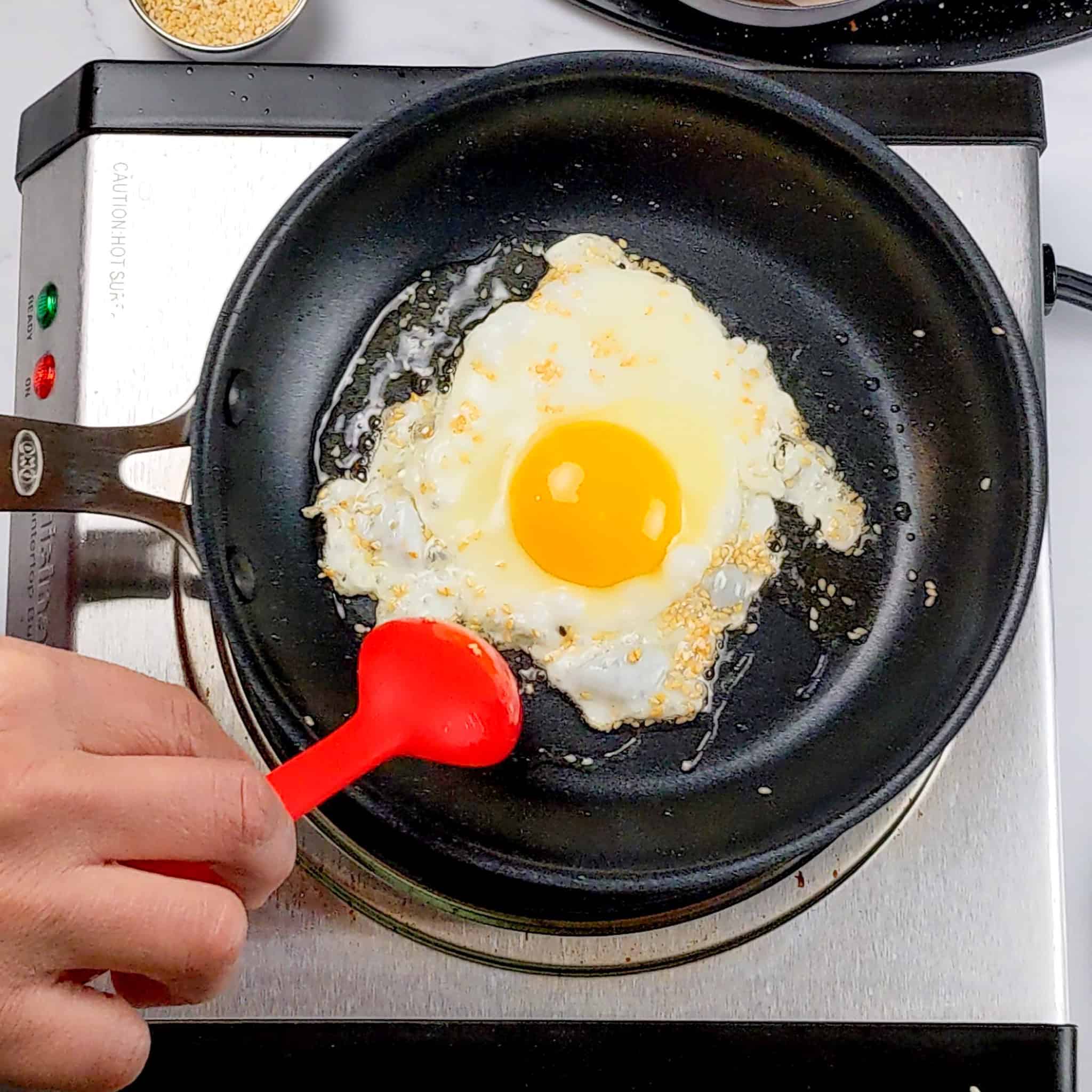 sesame seeds and oil being spooned over the fried sunny side egg with a silicone spoon made by GIR.