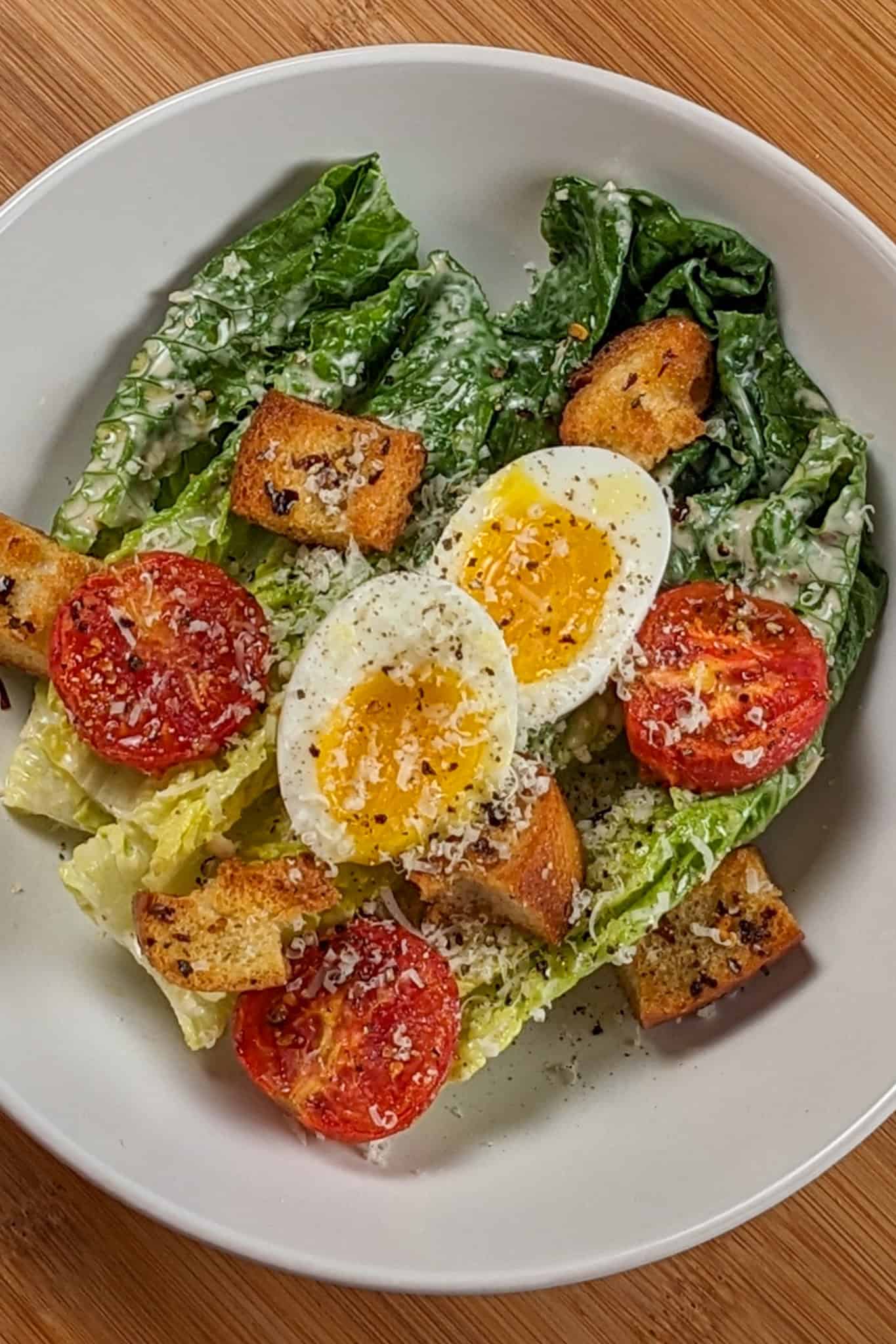 Caesar Salad with soft-boiled Egg, roasted campari Tomatoes and Chili-Infused Croutons garnished with freshly grated parmesan cheese and cracked black pepper in a wide rim bowl.