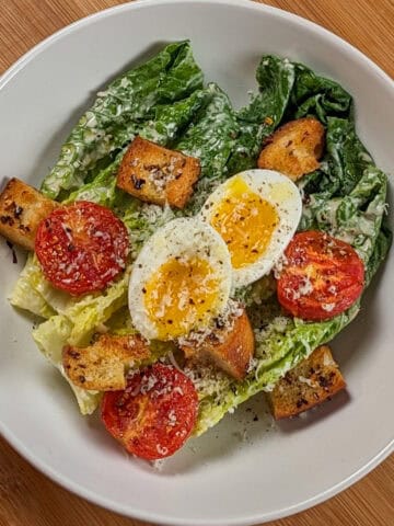Caesar Salad with soft-boiled Egg, roasted campari Tomatoes and Chili-Infused Croutons garnished with freshly grated parmesan cheese and cracked black pepper in a wide rim bowl.