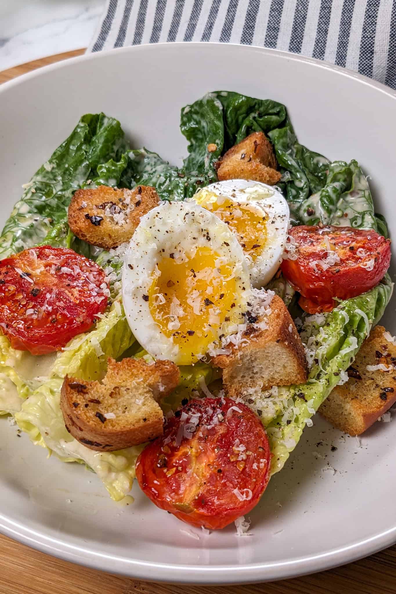 Caesar Salad with soft-boiled Egg, roasted campari Tomatoes and Chili-Infused Croutons garnished with freshly grated parmesan cheese and cracked black pepper in a wide rim bowl.