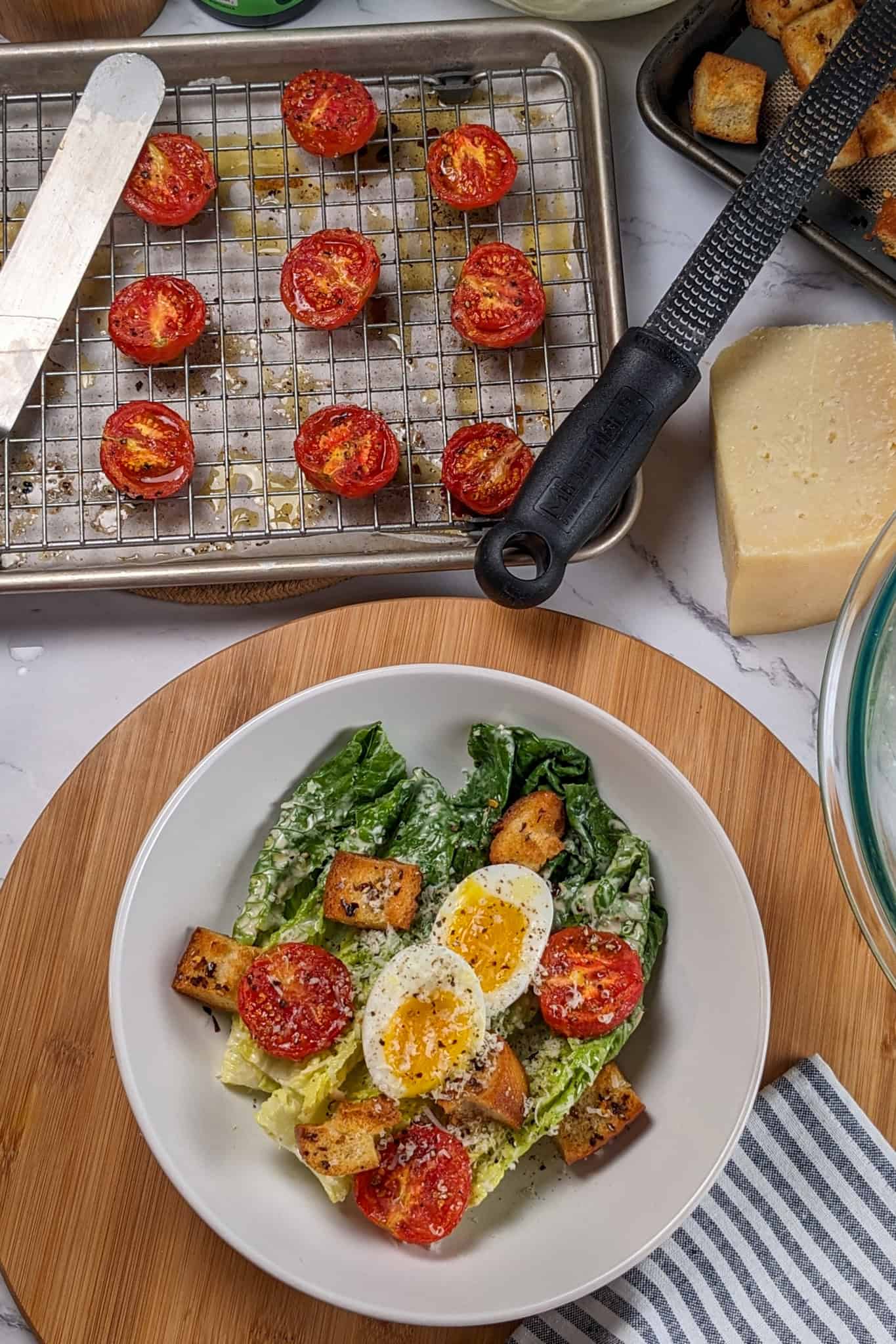 Caesar Salad with soft-boiled Egg, roasted campari Tomatoes and Chili-Infused Croutons garnished with freshly grated parmesan cheese and cracked black pepper in a wide rim bowl surrounded by a quarter sheet pan and rack with more roasted halved tomatoes, another sheet pan of croutons, parmesan wedge and a microplane.