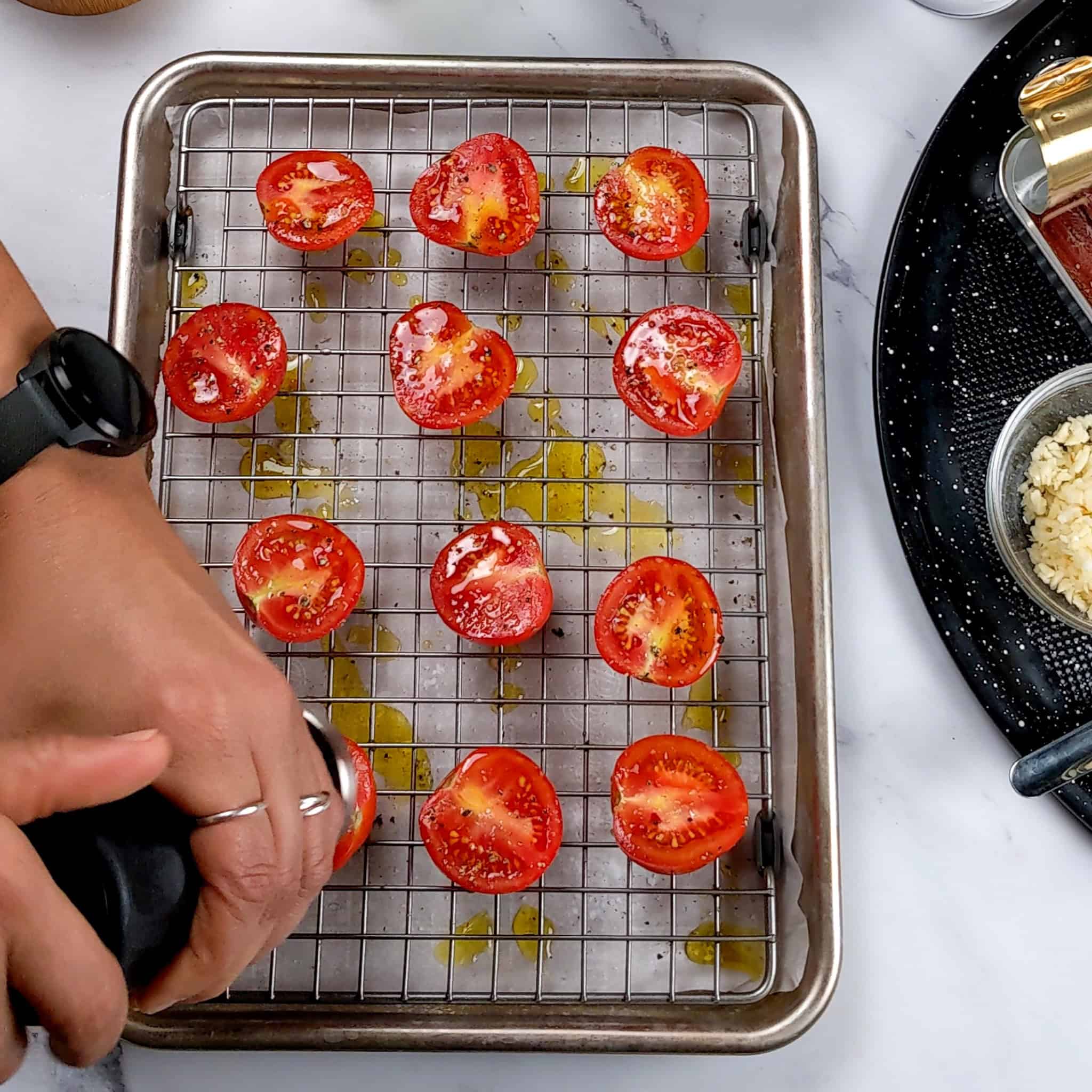 halved tomatoes evenly spread out on a sheet pan lined with parchment paper and a rack being seasoned with fresh cracked black pepper.