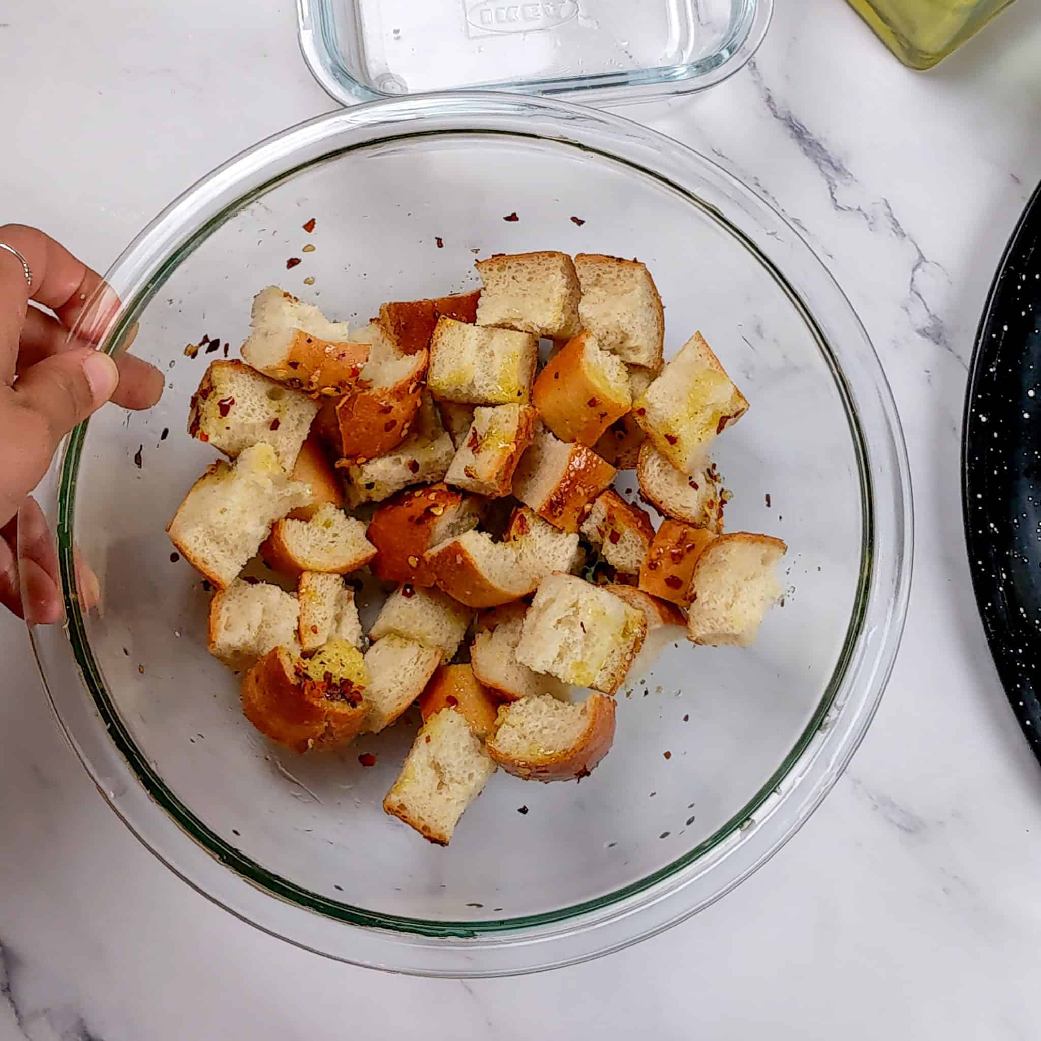 glass mixing bowl with bread chunks, olive oil, salt and pepper, and crushed red pepper chili flakes.