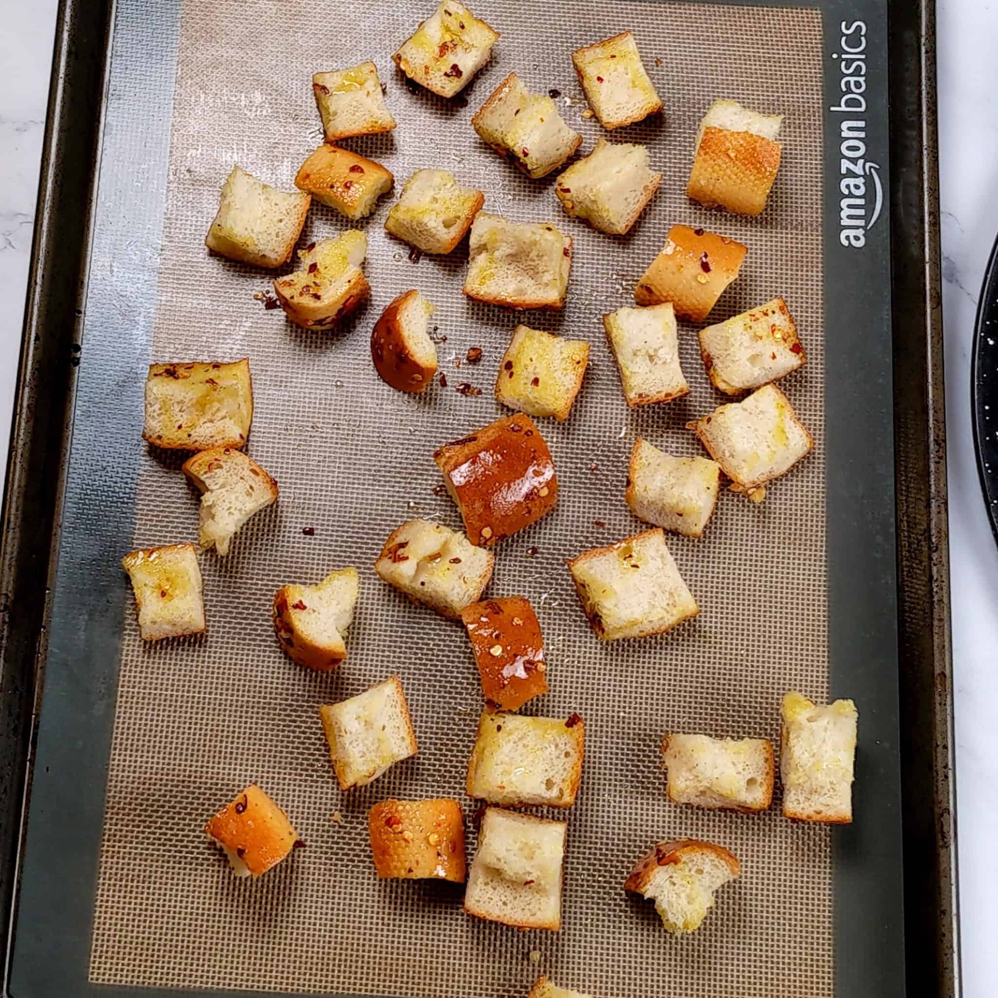 seasoned bread chunks with red pepper chili flakes and olive oil on a sheet pan lined with a silicone liner.