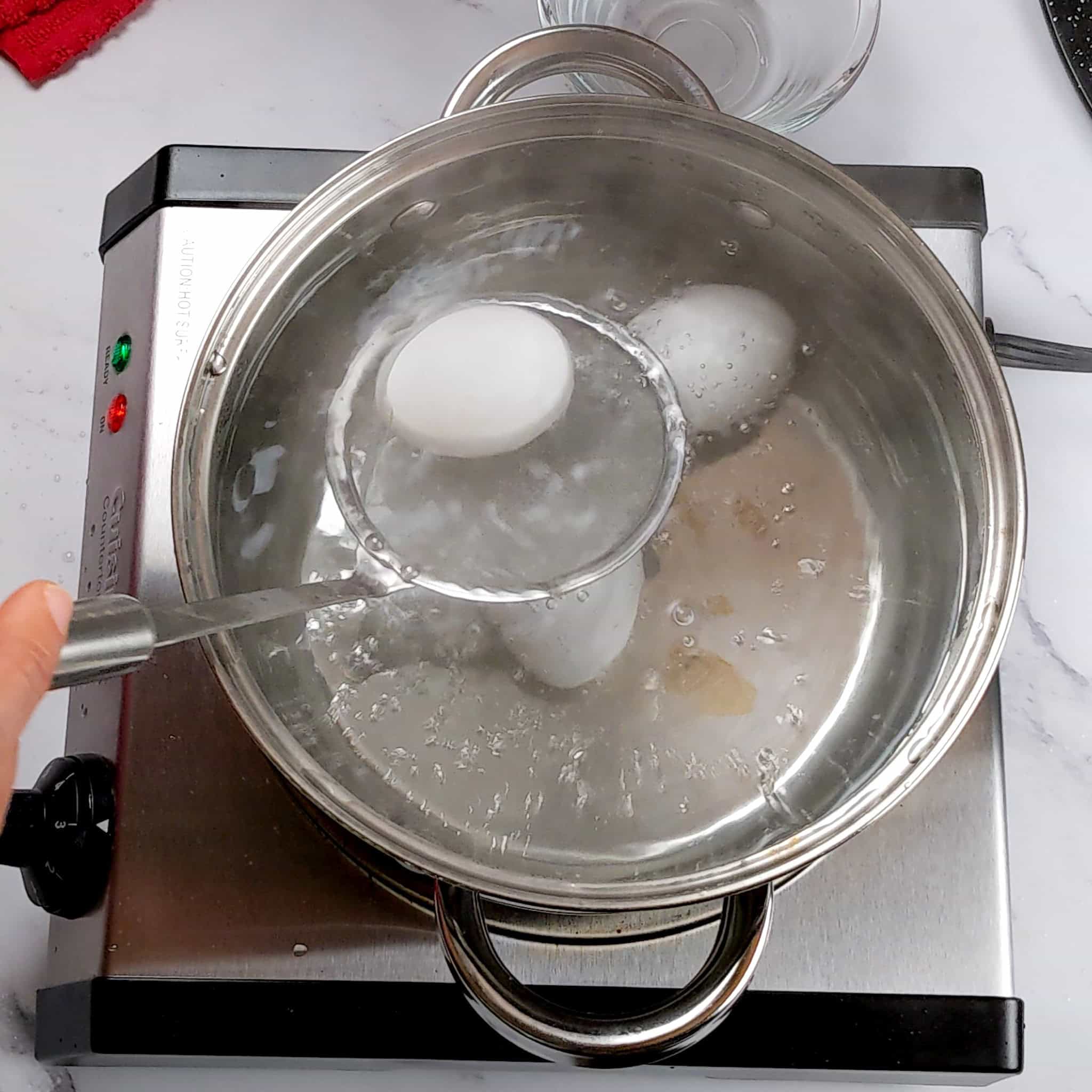 medium pot with boiling water with three eggs one being placed gently in with a skimmer.