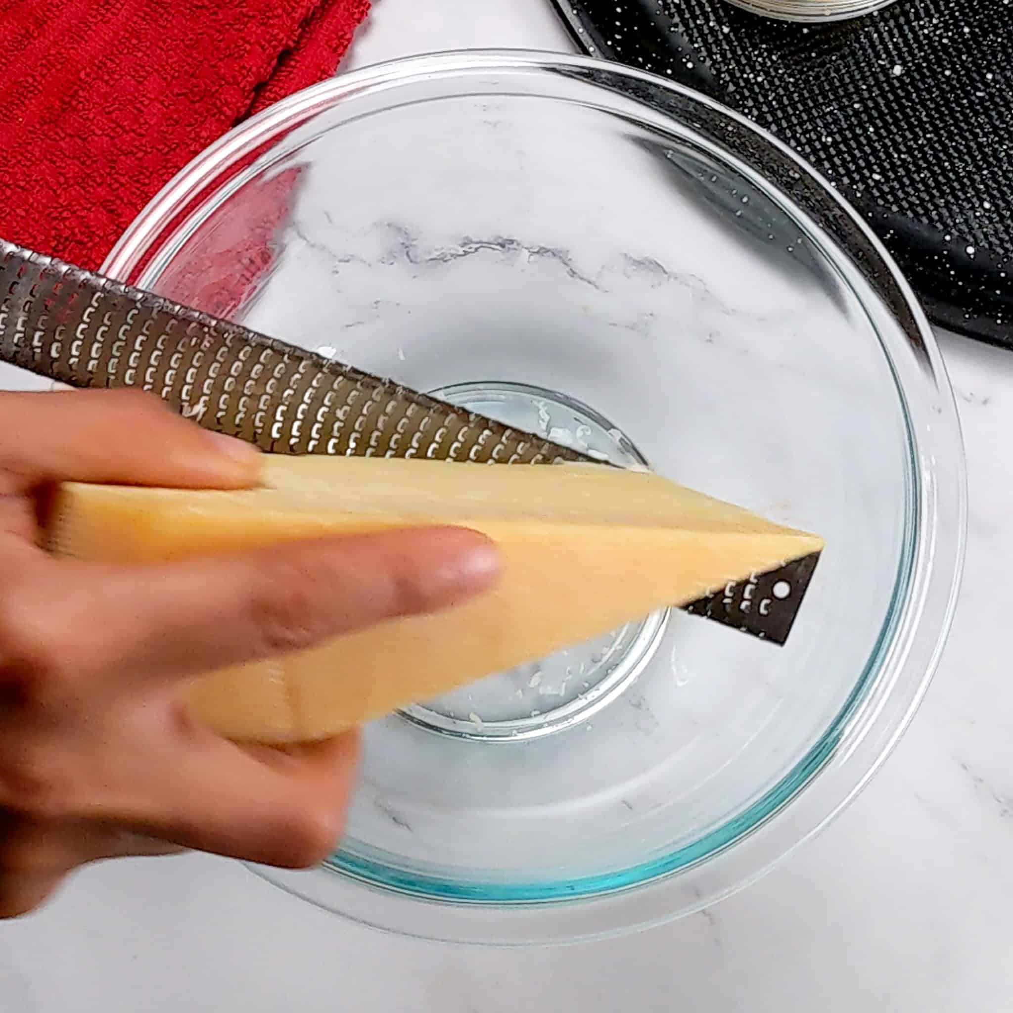 a parmesan cheese wedge being grated with a microplane over a glass mixing bowl.