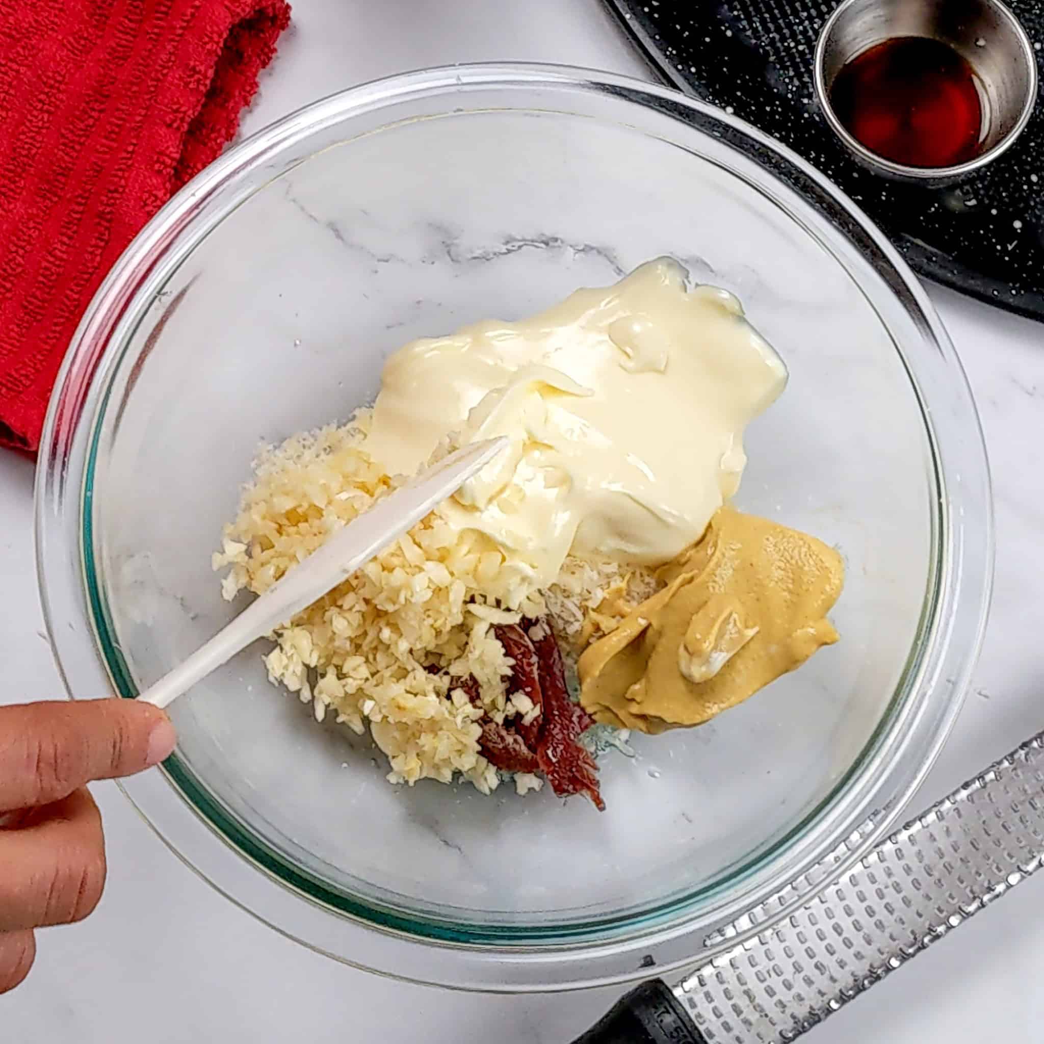 glass mixing bowl with mayonnaise, chopped garlic, anchovies, Dijon mustard being mixed with a mini silicone spatula.