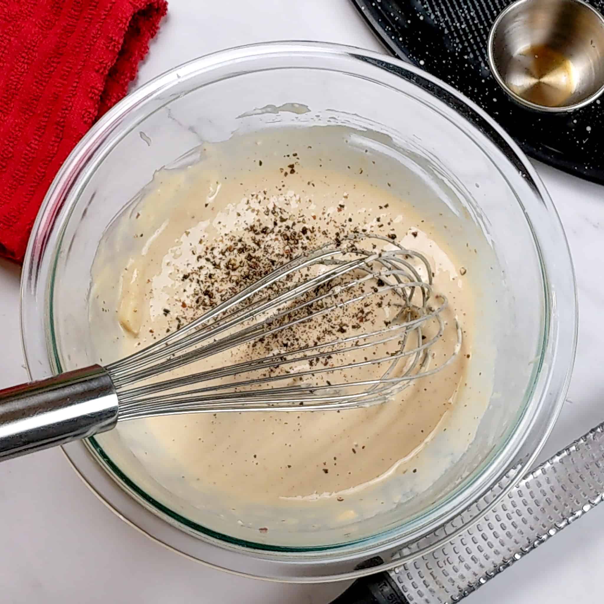 freshly made Ceasar salad dressing in a glass mixing bowl with a whisk sitting inside and topped with freshly cracked black pepper.