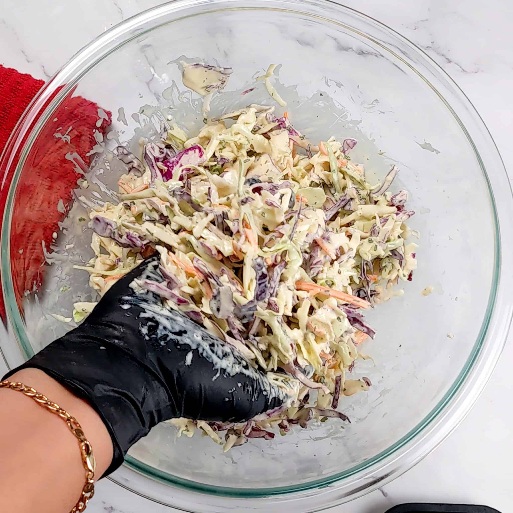 homemade coleslaw in a glass mixing bowl being mixed with a gloved hand.