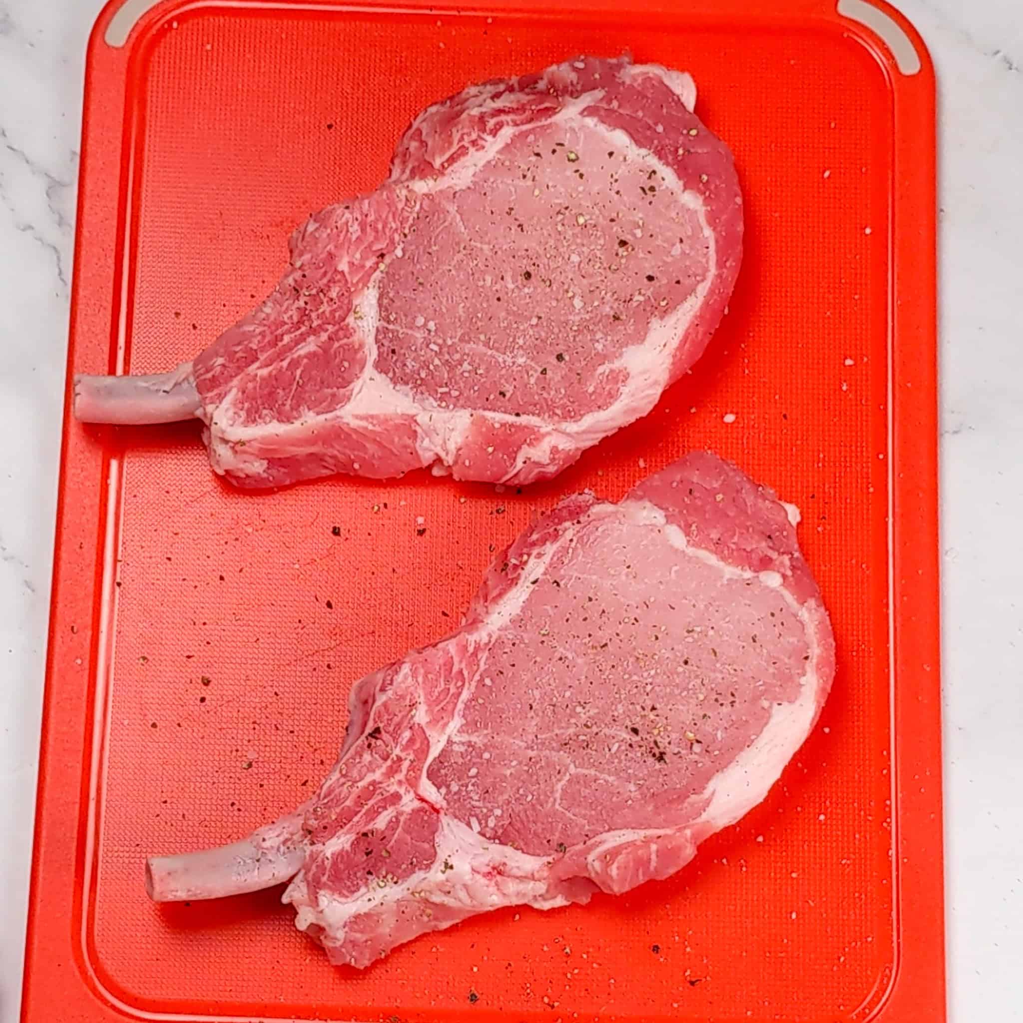 salt and pepper seasoned rib pork chops on a color coded cutting board for raw meat.