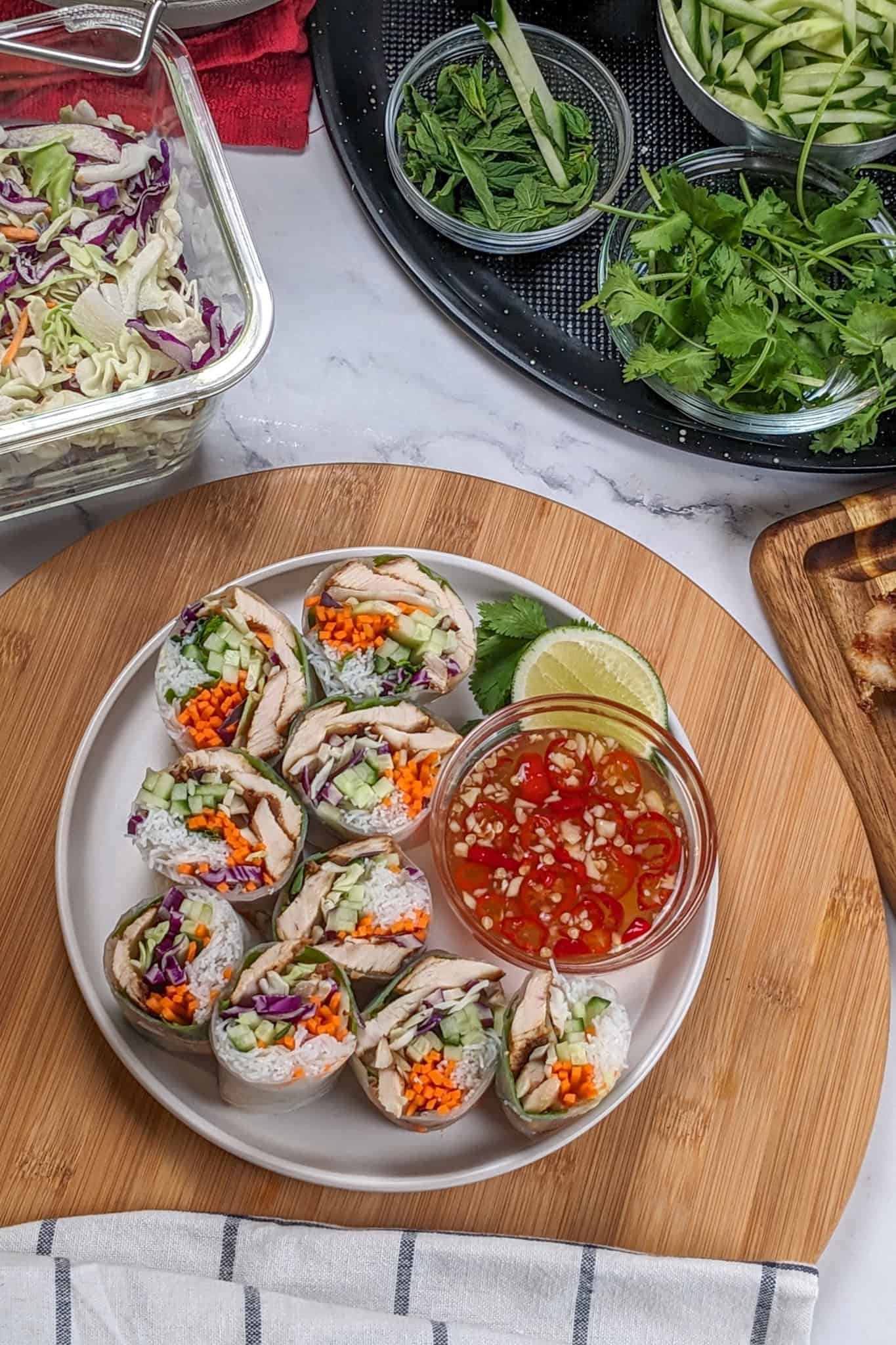 sliced lemongrass chicken, cabbage, rice vermicelli spring roll with nouc cham sauce on side all on a round flat plate surrounded by glass container of shredded tri-color slaw, and other ingredients.