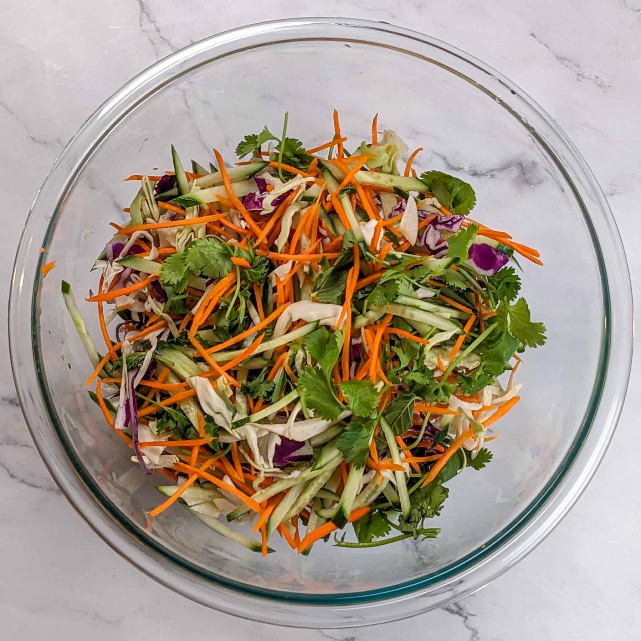 shredded green and red cabbage, carrots, cucumber, cilantro leaves, and mint leaves in a glass mixing bowl.