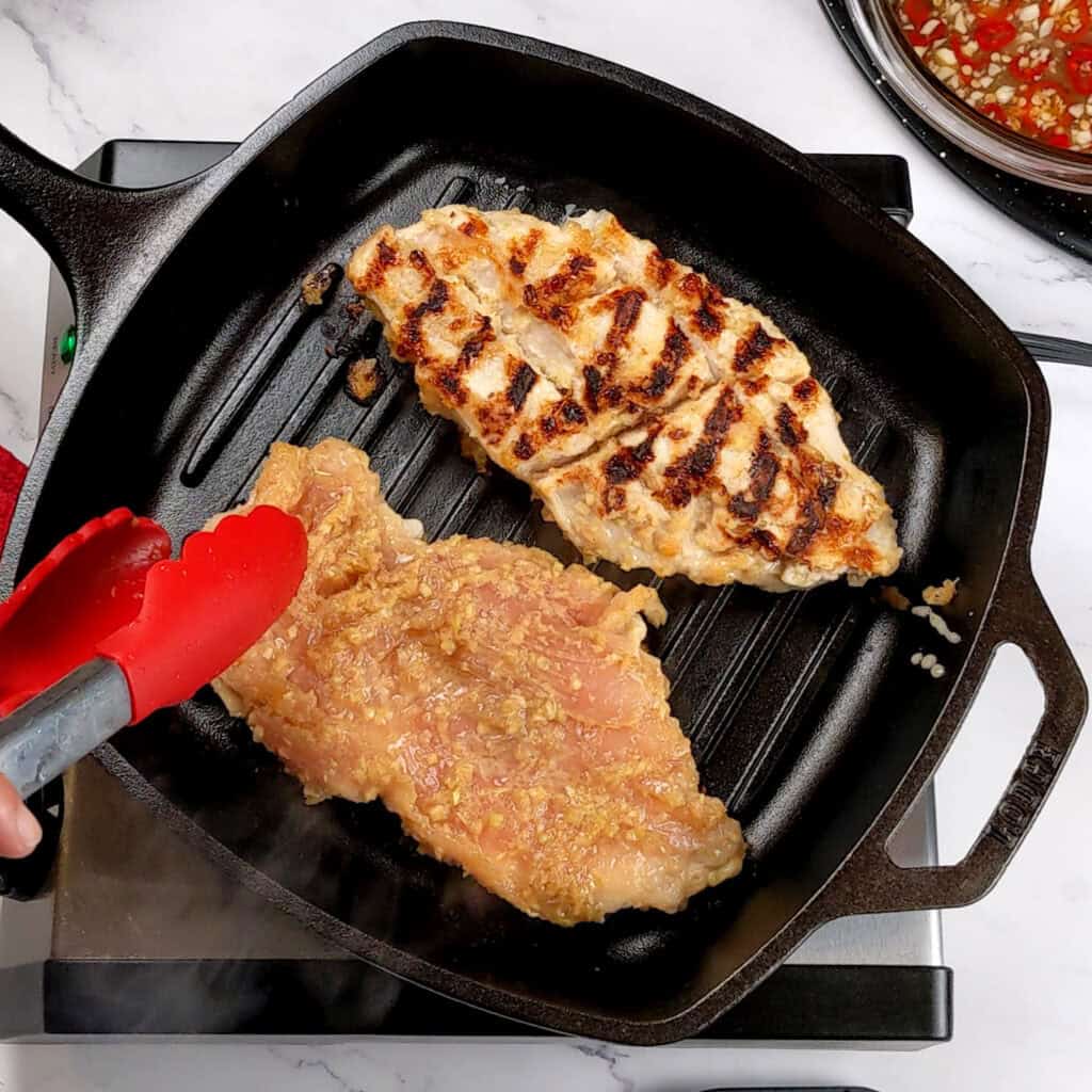 two grilled lemongrass chicken breast with grill marks on one and another about to be flipped with a kitchenaid silicone spatula in a grill pan.