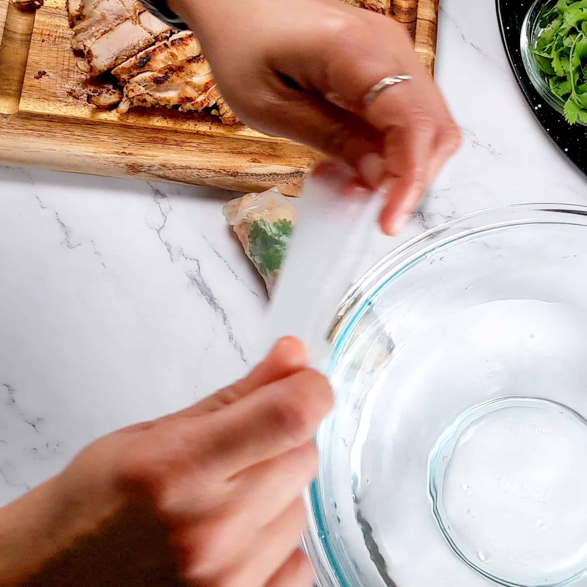 soaked rice paper pulled out of a glass pyrex mixing bowl with water by two hands.