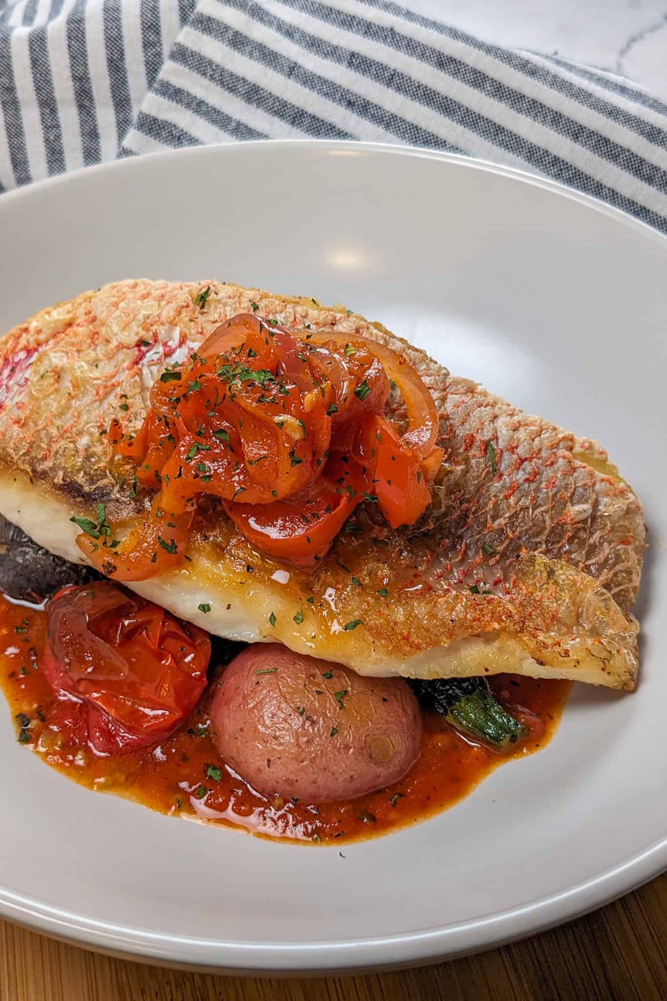 Pan seared snapper toped with stewed onion and red bell pepper garnish on a bed of roasted baby vegetables on spicy creole sauce in a wide rim round bowl.