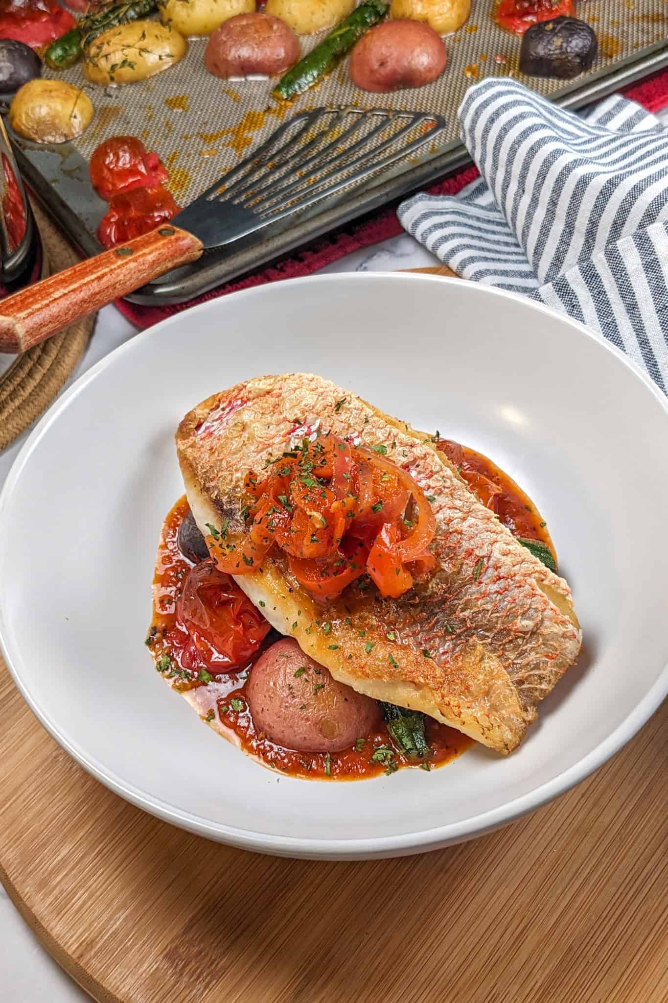 Pan seared snapper toped with stewed onion and red bell pepper garnish on a bed of roasted baby vegetables on spicy creole sauce in a wide rim round bowl surrounded by more roasted vegetables on a sheet pan lined with a silicone liner with a fish spatula laying inside.