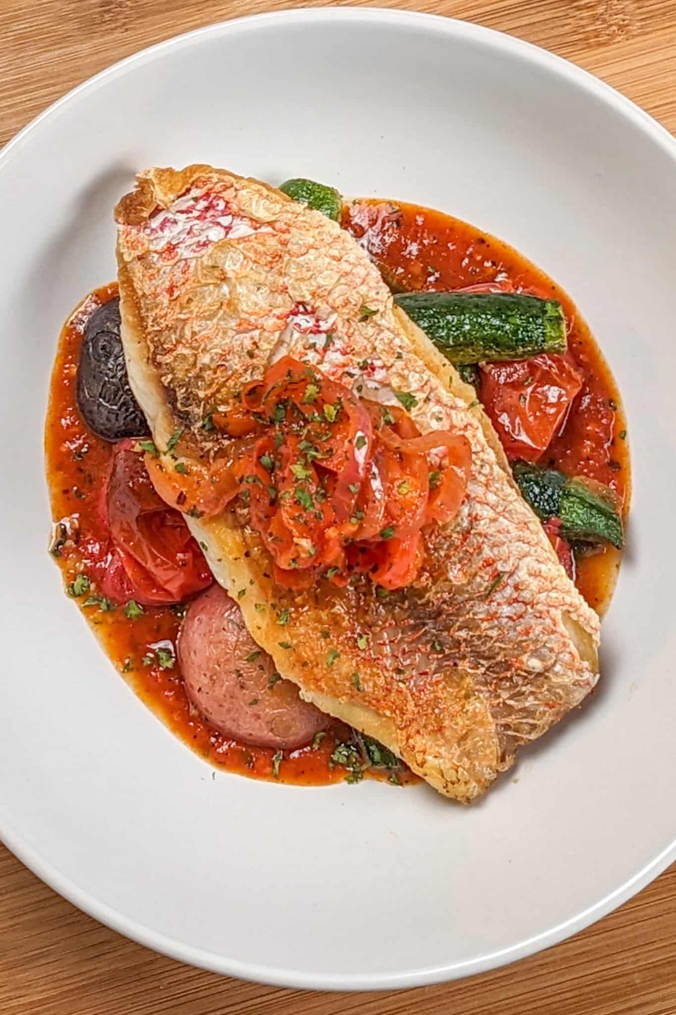 Pan seared snapper toped with stewed onion and red bell pepper garnish on a bed of roasted baby vegetables on spicy creole sauce in a wide rim round bowl.