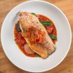 Pan seared snapper toped with stewed onion and red bell pepper garnish on a bed of roasted baby vegetables on spicy creole sauce in a wide rim round bowl.