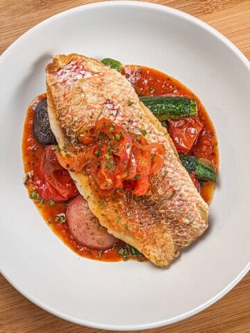 Pan seared snapper toped with stewed onion and red bell pepper garnish on a bed of roasted baby vegetables on spicy creole sauce in a wide rim round bowl.