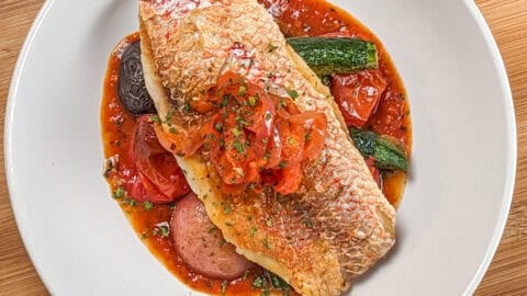 Pan seared snapper toped with stewed onion and red bell pepper garnish on a bed of roasted baby vegetables on spicy creole sauce in a wide rim round bowl.