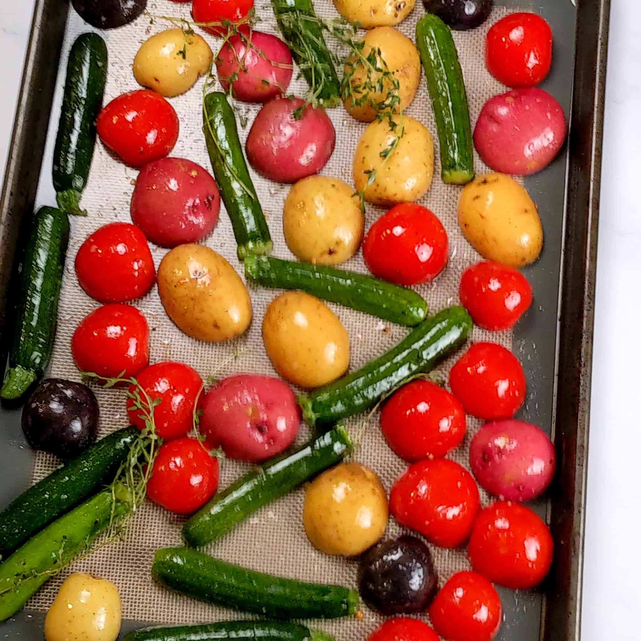 olive oil, fresh thyme, salt and cracked black pepper seasoned baby vegetables of halved baby zucchini, different color baby potatoes, and campari tomatoes on a silicone lined sheet pan.