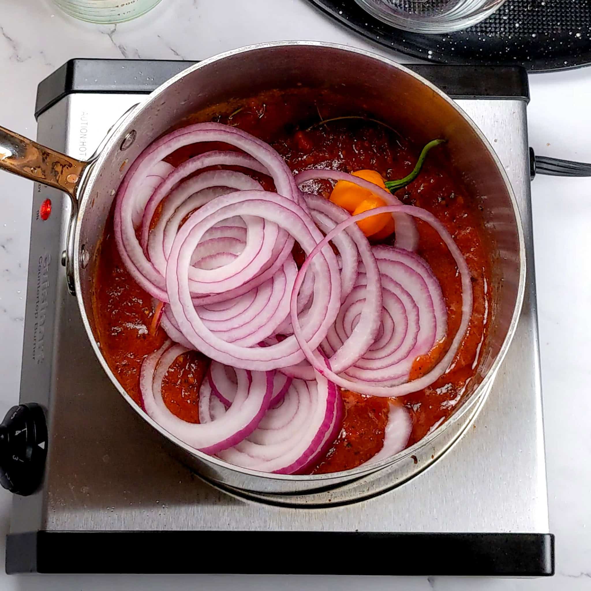 spicy creole sauce topped with a pierced whole scotch bonnet pepper and sliced red onion rings in an all-clad sauce pot.