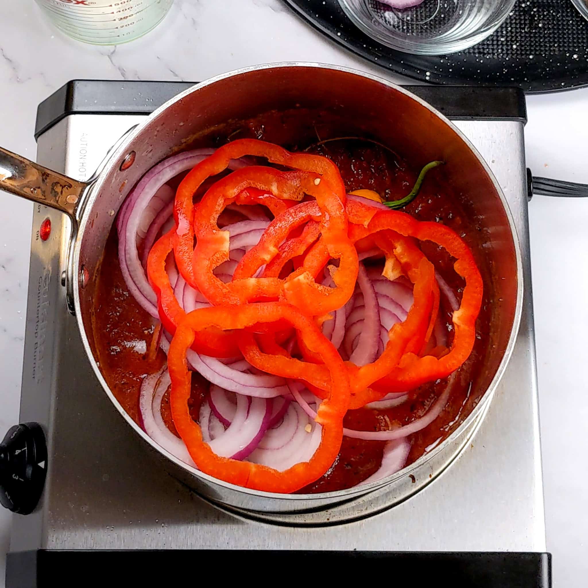 spicy creole sauce topped with a pierced whole scotch bonnet pepper and sliced red onion rings and red bell pepper sliced rings in an all-clad sauce pot.