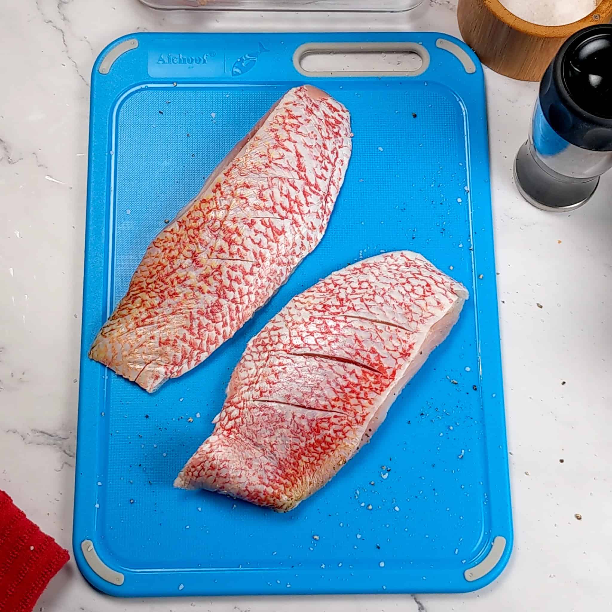 slitted salt and pepper seasoned red snapper filets on a color coded plastic cutting board for raw fish .