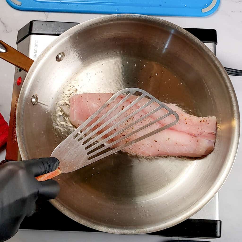 fish spatula pressing down on a snapper filet skin side down frying in an stainless steel all clad frying pan.