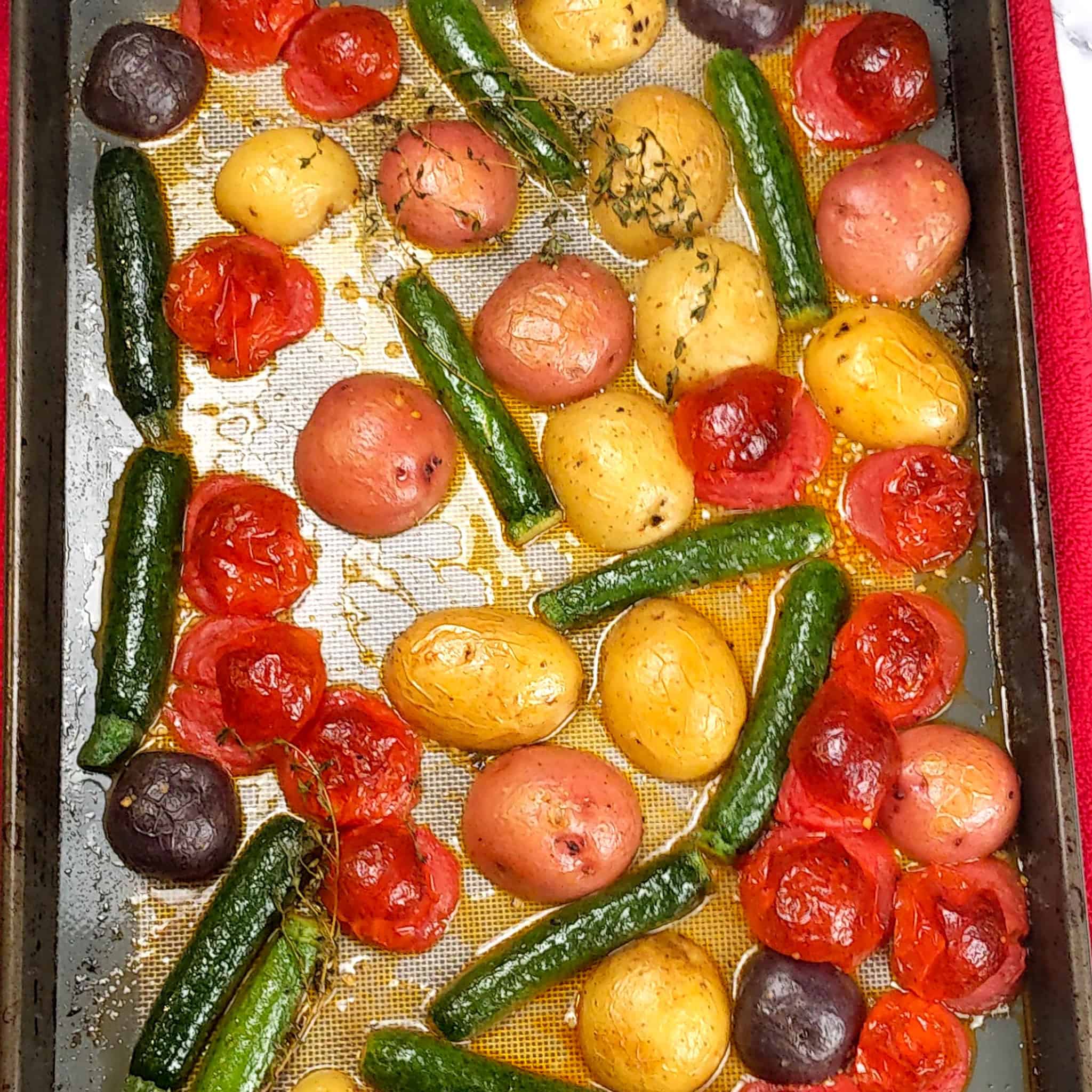 Roasted olive oil, fresh thyme, salt and cracked black pepper seasoned baby vegetables of halved baby zucchini, different color baby potatoes, and campari tomatoes on a silicone lined sheet pan.