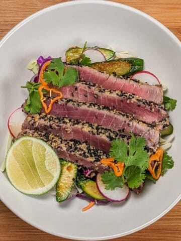 Sesame crusted ahi tuna on a bed of cabbage slaw, carrots, radishes, and zucchini with soy-sesame dressing and garnished with fresh cut lime, cilantro leaves and habanero slices on a wide rim bowl.