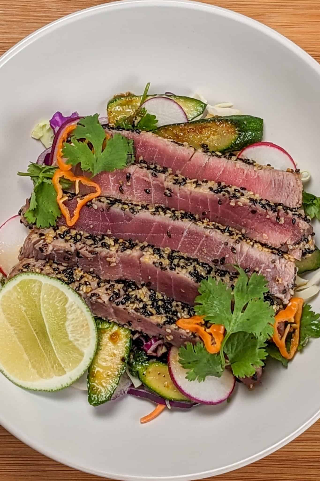 Sesame crusted ahi tuna on a bed of cabbage slaw, carrots, radishes, and zucchini with soy-sesame dressing and garnished with fresh cut lime, cilantro leaves and habanero slices on a wide rim bowl.