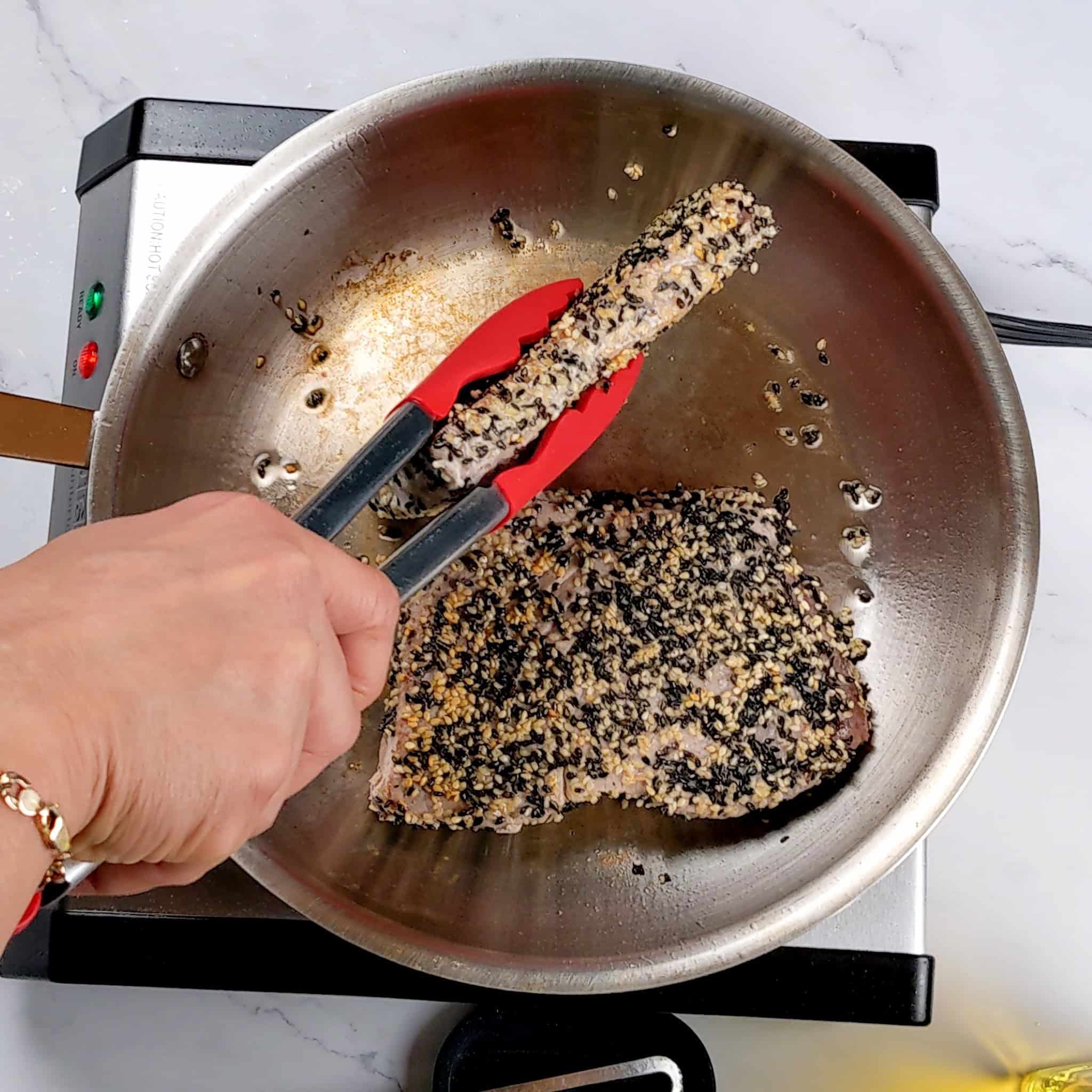 sesame seed crusted ahi tuna being seared in a skillet with one being held on its side with silicone kitchenaid tongs.