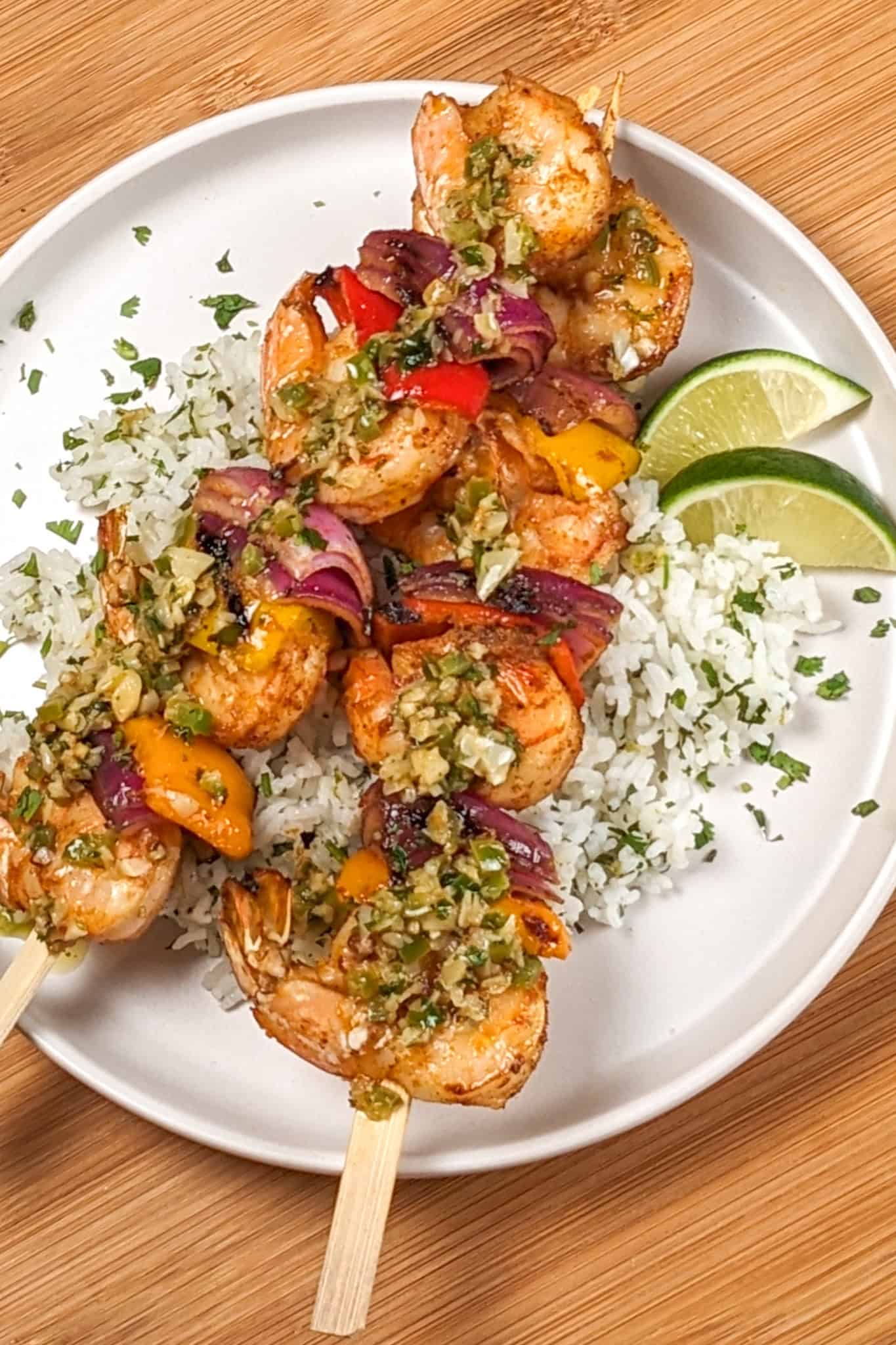 two Grilled Shrimp skewers with Garlic Jalapeno Butter and Cilantro Lime Rice on a flat round plate garnished with lime wedges and chopped cilantro.