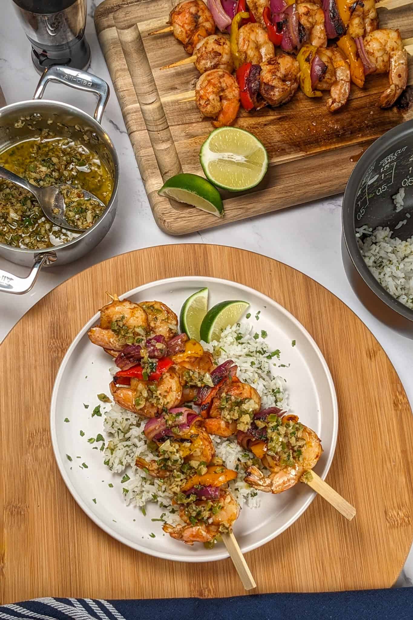 two Grilled Shrimp skewers with Garlic Jalapeno Butter and Cilantro Lime Rice on a flat round plate garnished with lime wedges and chopped cilantro surrounded by a mini all-clad sauce pot, and more shrimp skewers on a wooden cutting board.