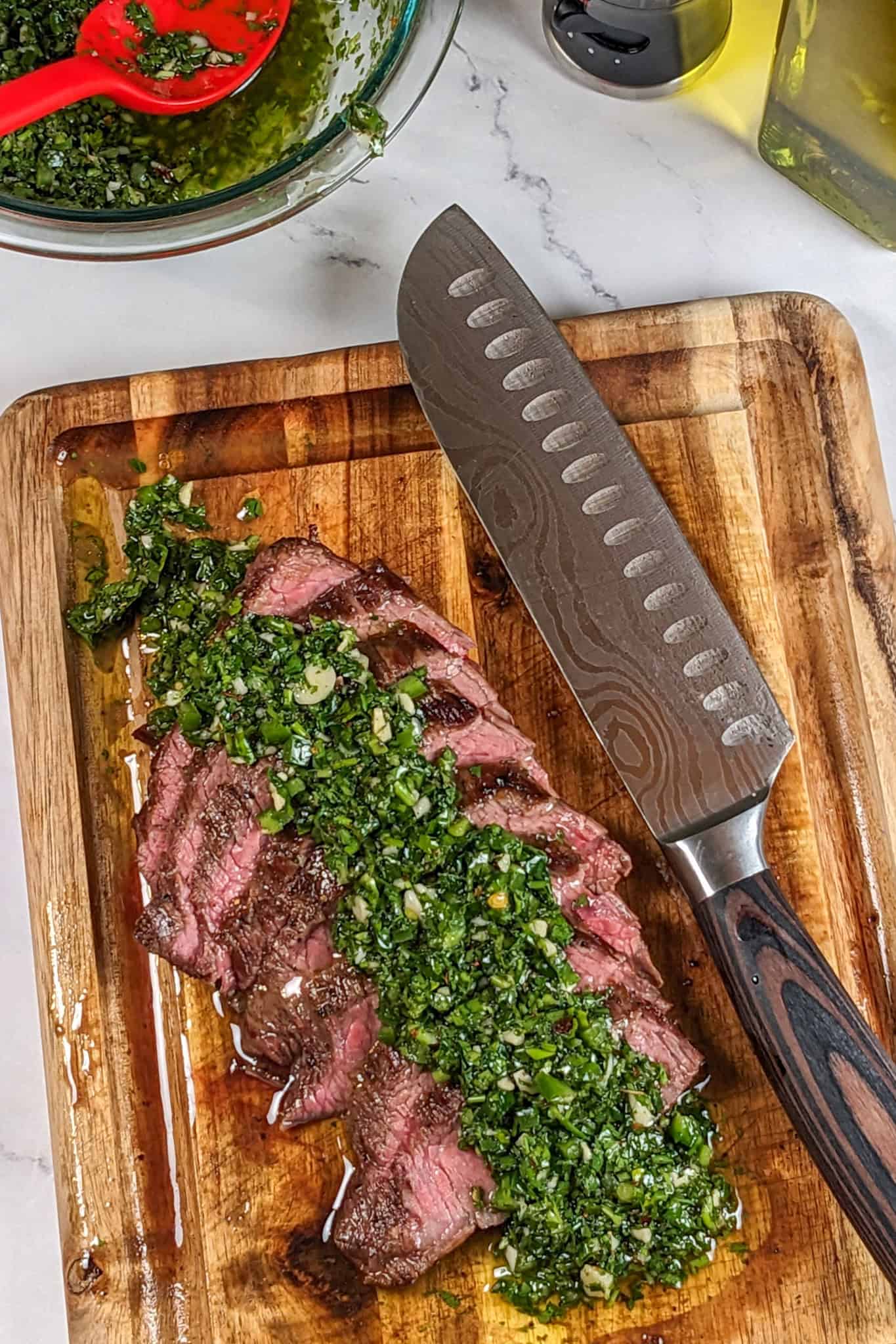 medium rare thinly sliced pan-grilled flank steak topped with spicy chimichurri sauce cascading on a mini wooden cutting board with a knife laying next to it.