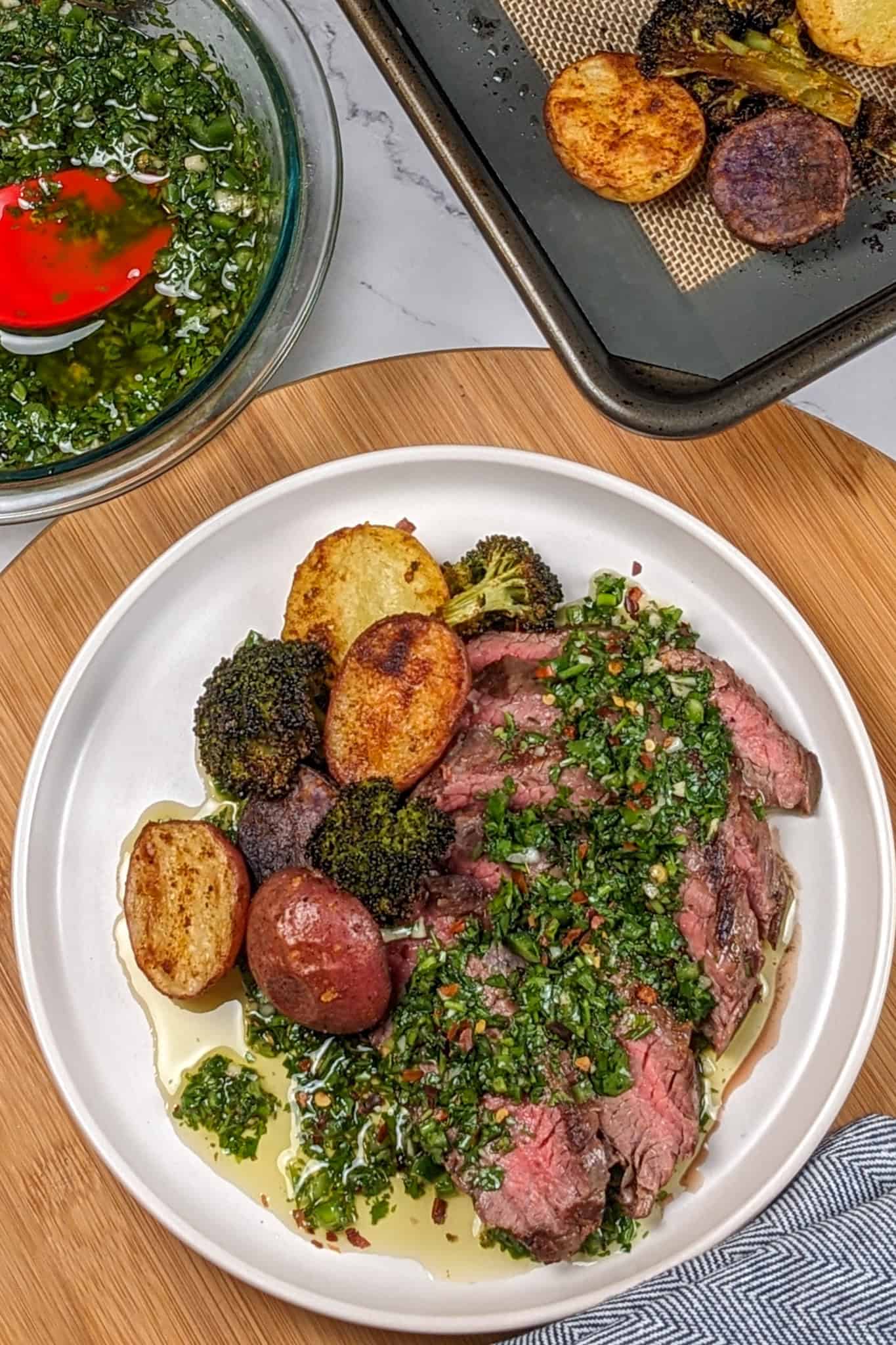 Grilled Steak with Spicy jalapeno Chimichurri sauce with a medley of roasted Baby Potatoes and Broccoli florets surrounded by chimichurri in a glass mixing bowl and a sheet pan lined with silicone liner with roasted baby potatoes and broccoli.