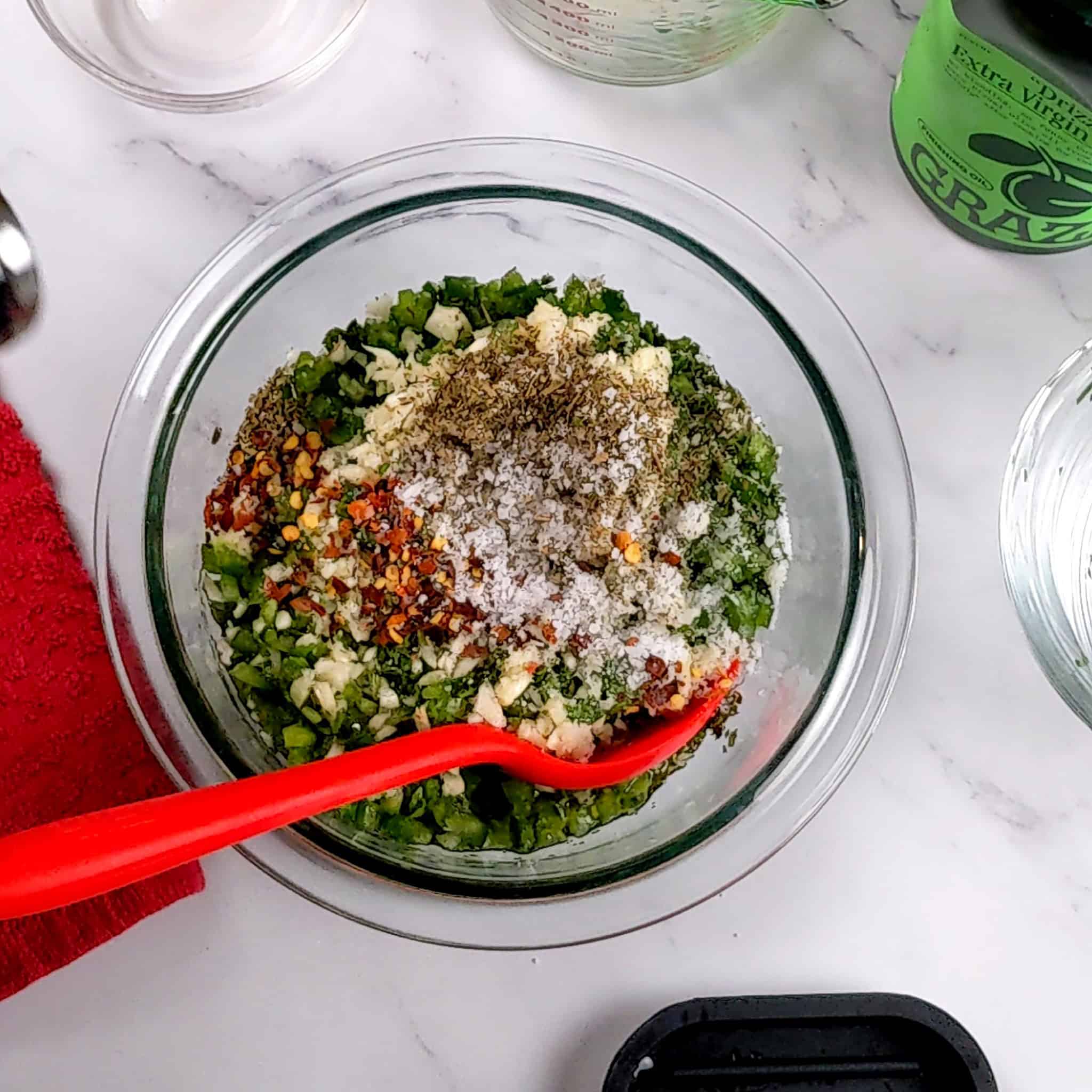 seasoning of ground black pepper, chopped garlic, red pepper chili flakes and kosher salt to chimichurri in a glass mixing bowl with a silicone spoon.