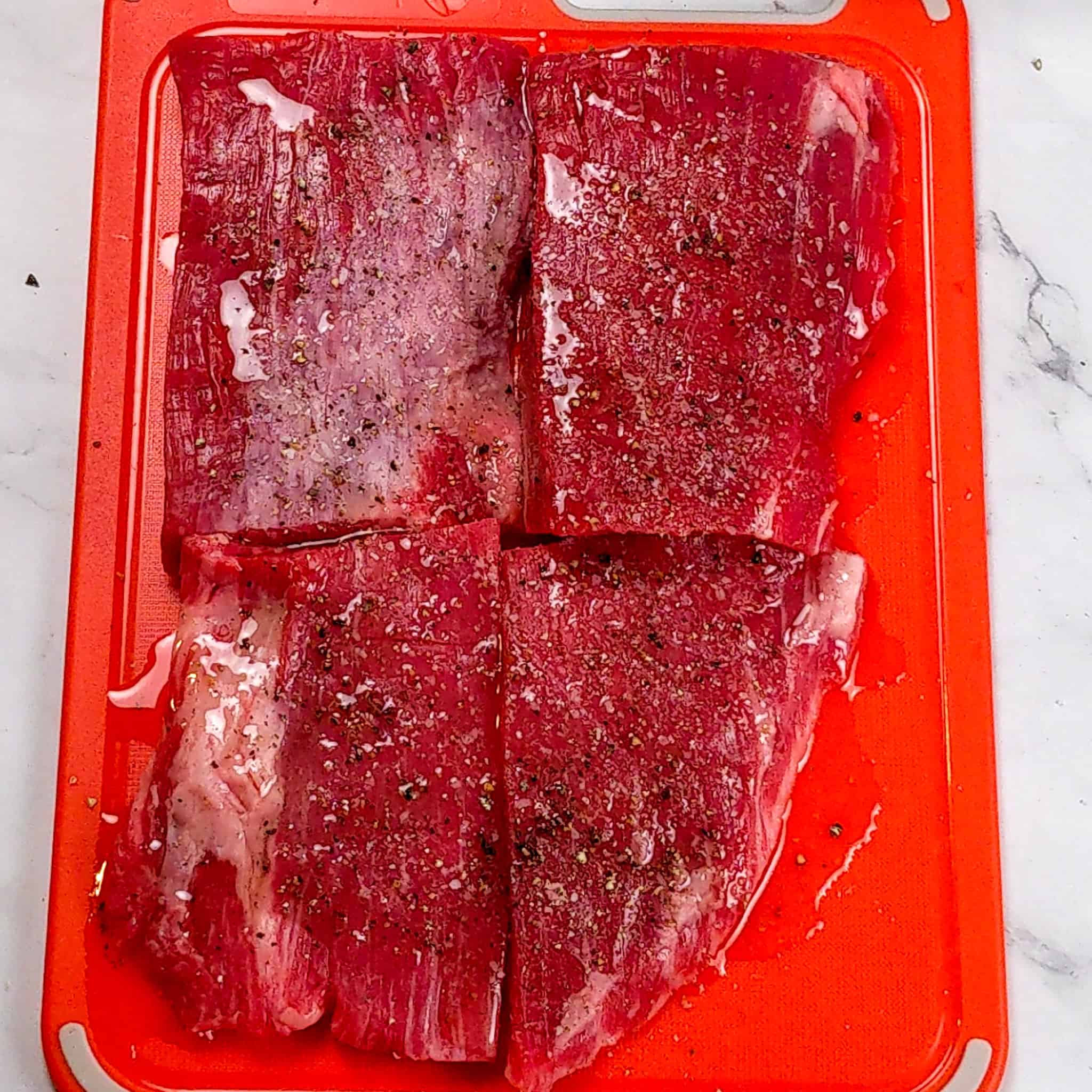 olive oil, ground black pepper and salted seasoned portioned flank steak on a raw meat coded plastic cutting board.